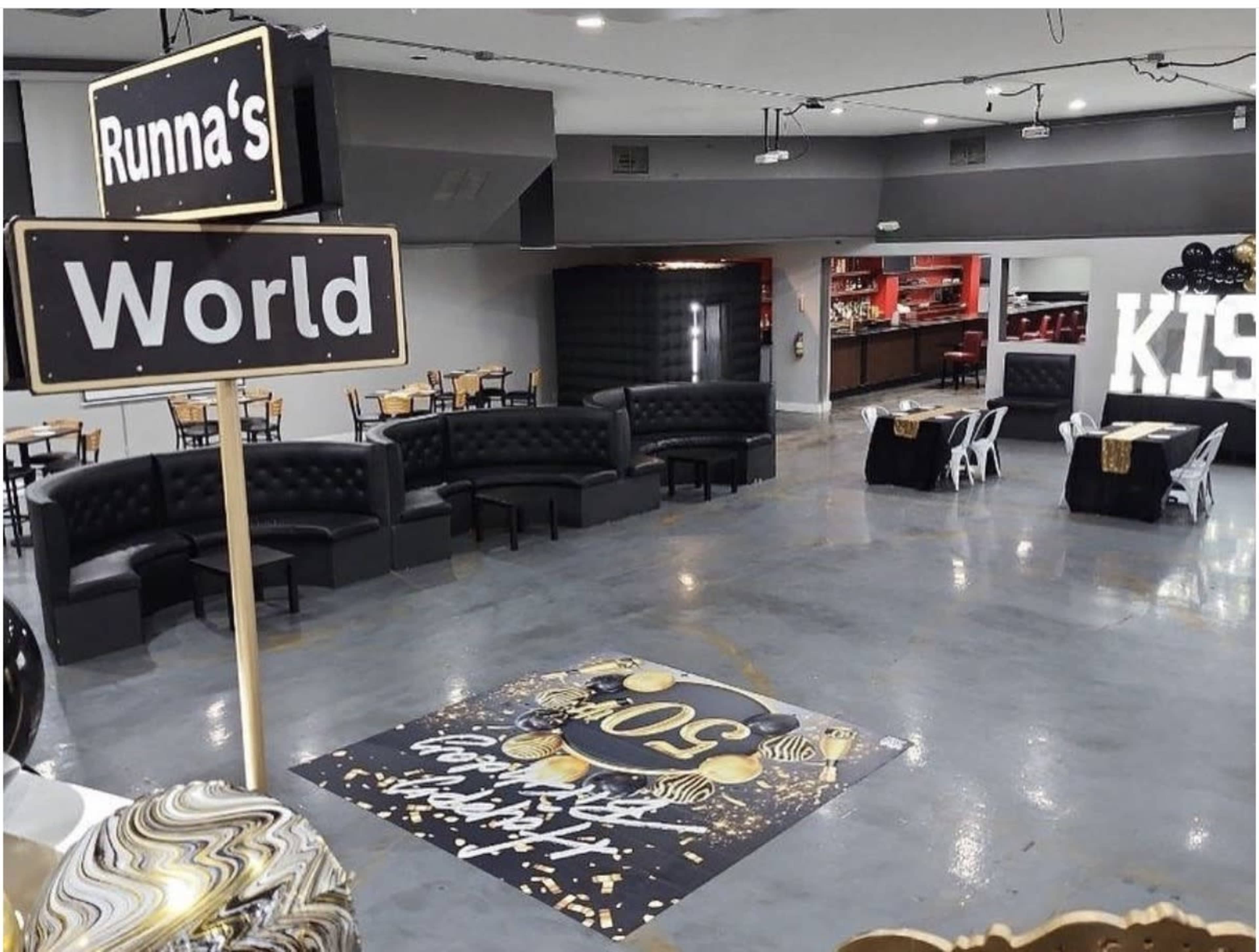 The image shows a spacious event venue with black leather seating arrangements, a decorative floor design celebrating a milestone, and a festive atmosphere indicated by balloons and signage.