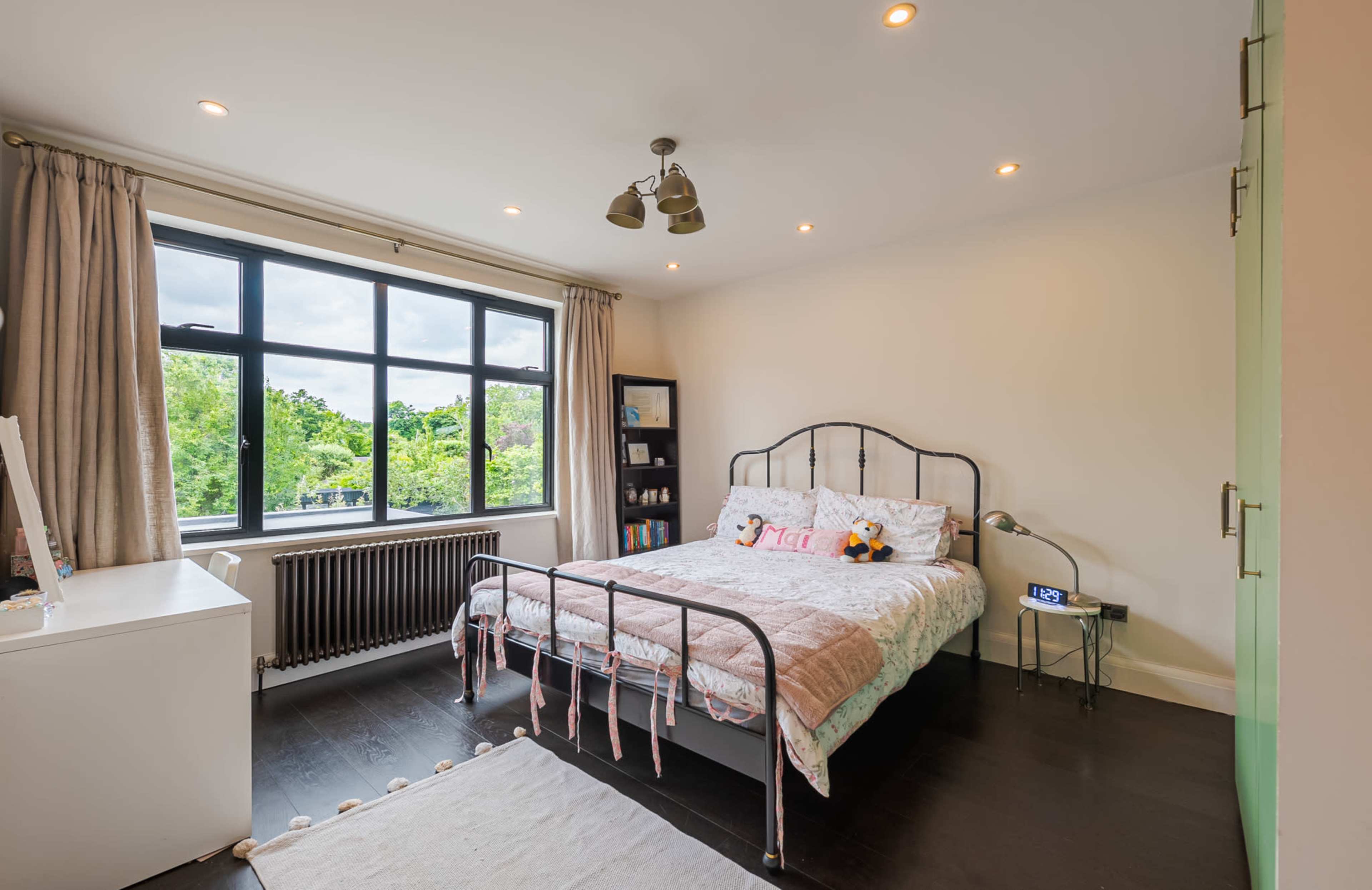 A well-lit bedroom features a bed with decorative pillows, large windows overlooking greenery, and a modern decor style.