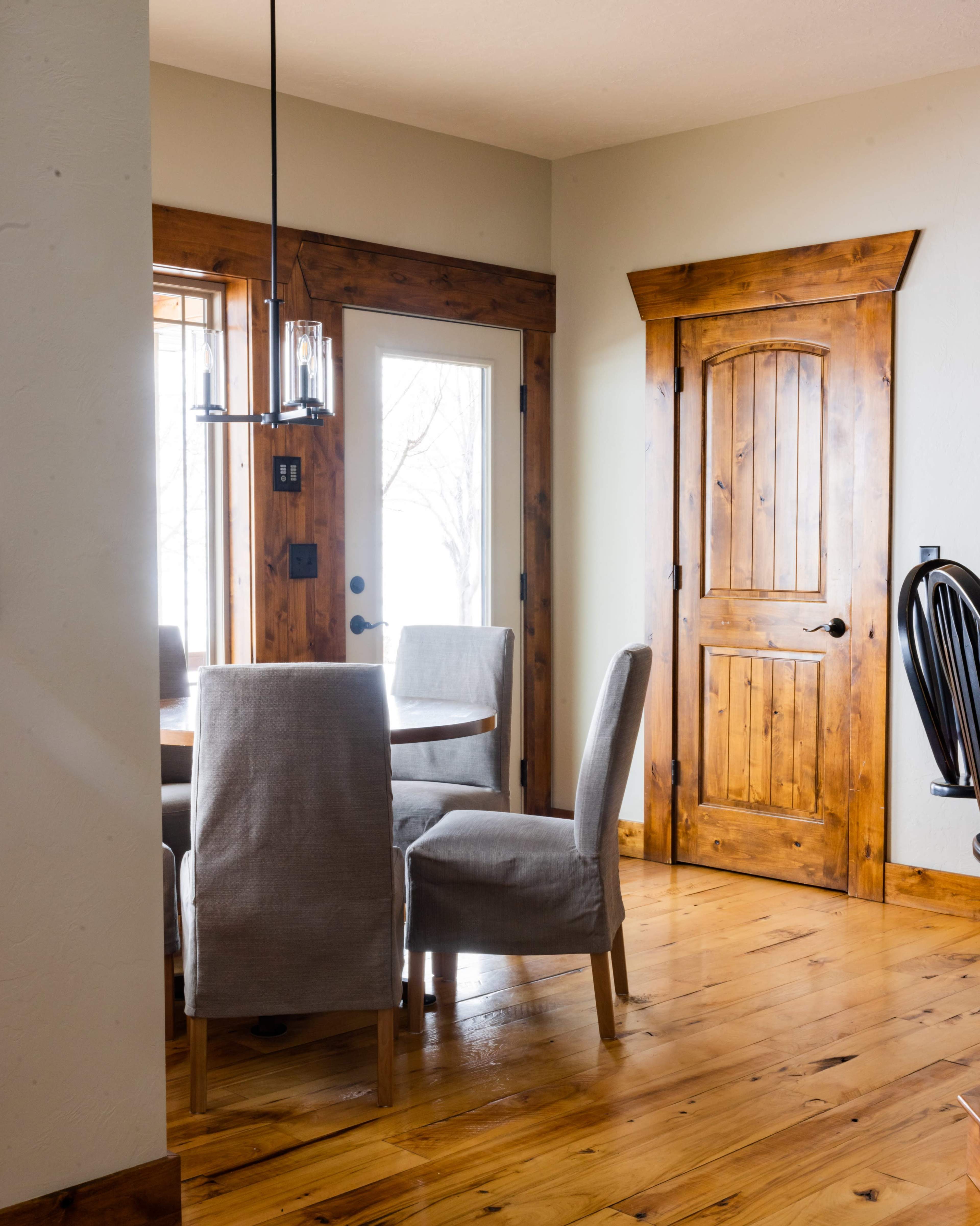 A dining area with a round table surrounded by four upholstered chairs, a wooden door leading outside, and a door to another room, all set on polished hardwood flooring.