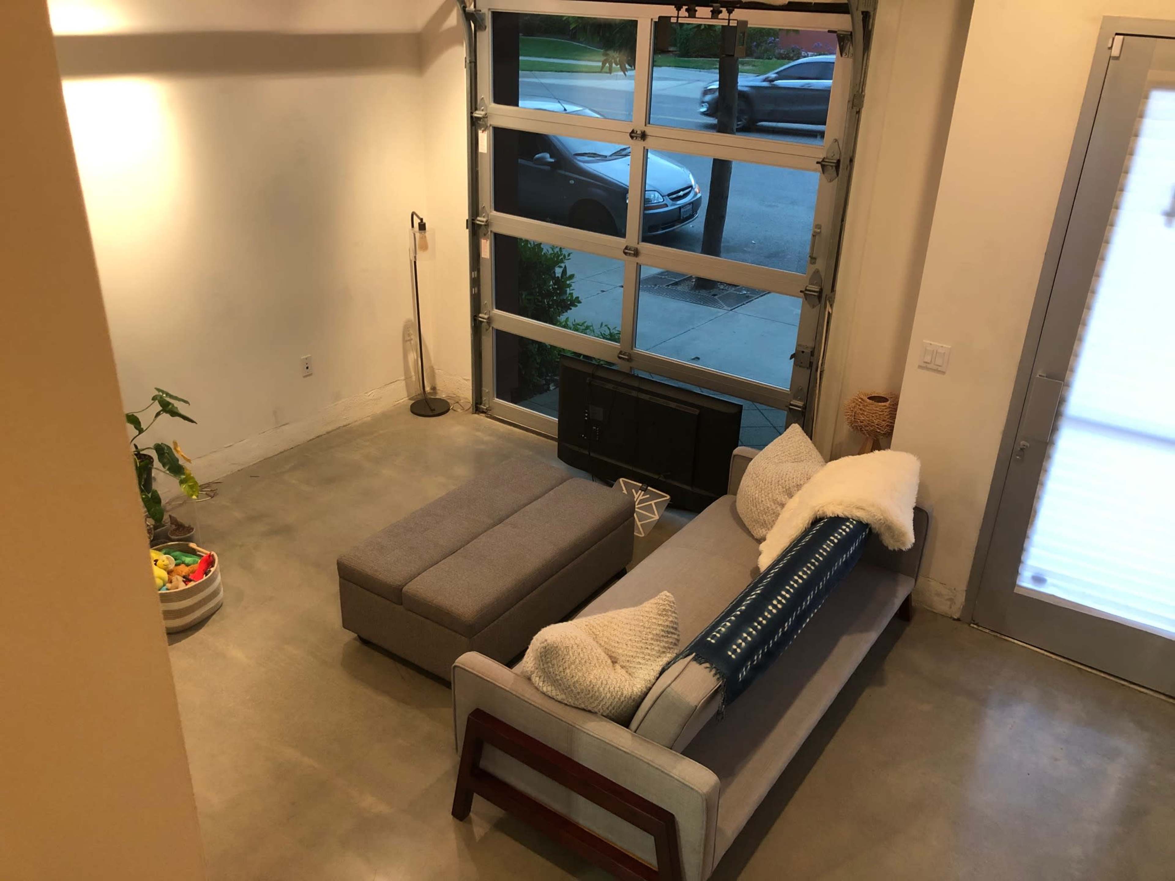 The image shows a minimalist living space with a gray sofa and ottoman, a floor lamp, and a large garage door opening to the street.