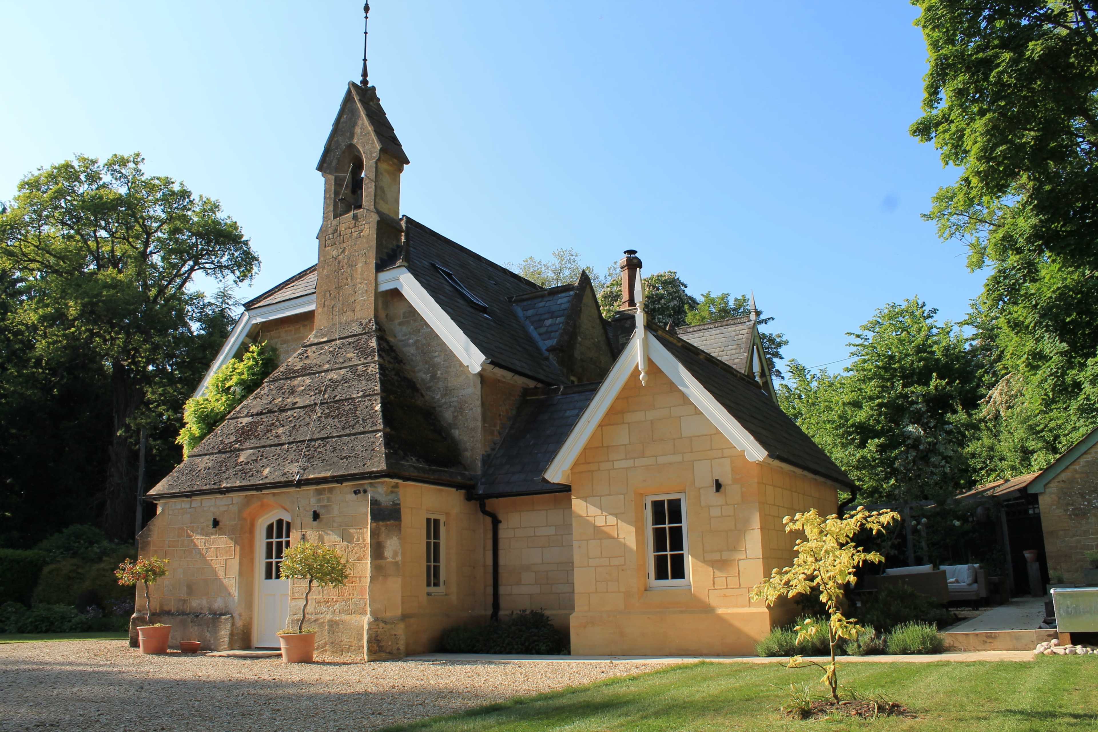 A stone cottage with a peaked roof and a small tower stands among lush greenery and a gravel driveway.