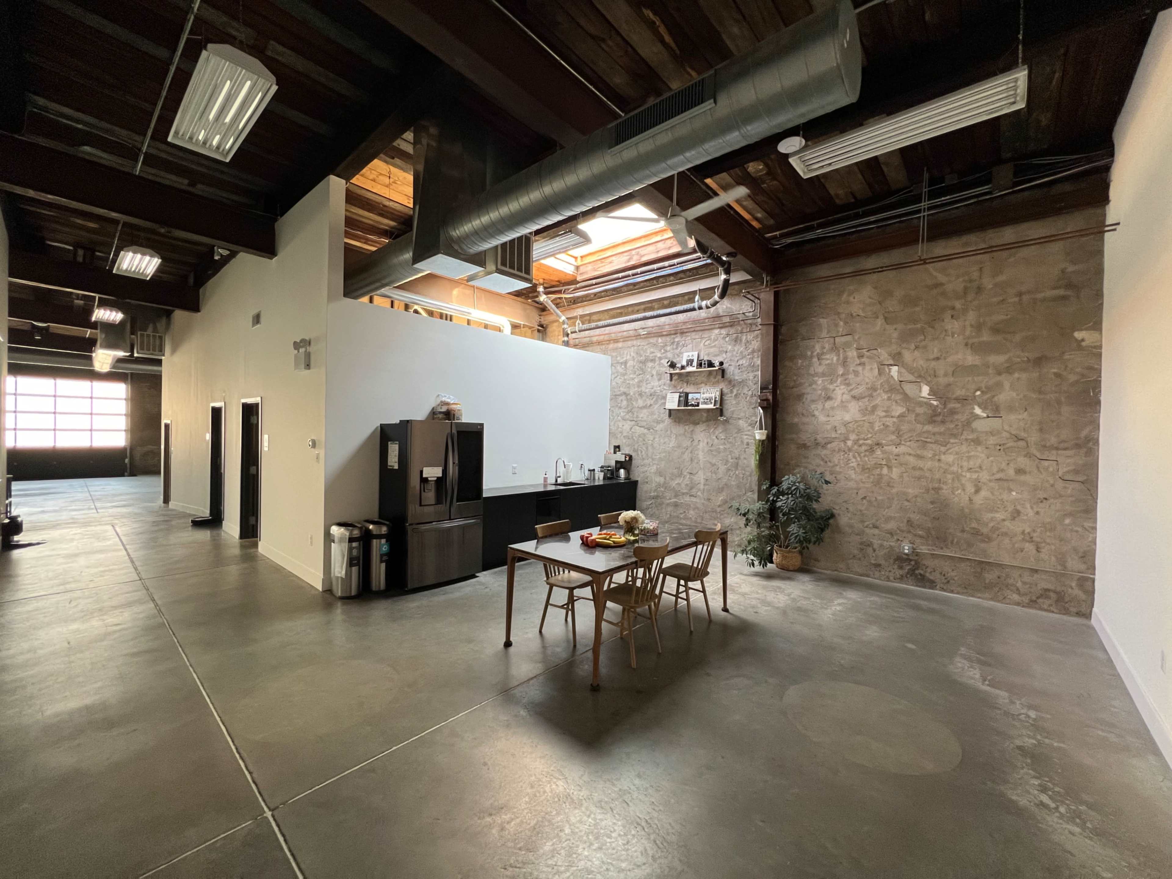 A spacious, industrial-style kitchen area featuring a black refrigerator, a wooden dining table, and a potted plant, with exposed brick walls and large windows overhead.