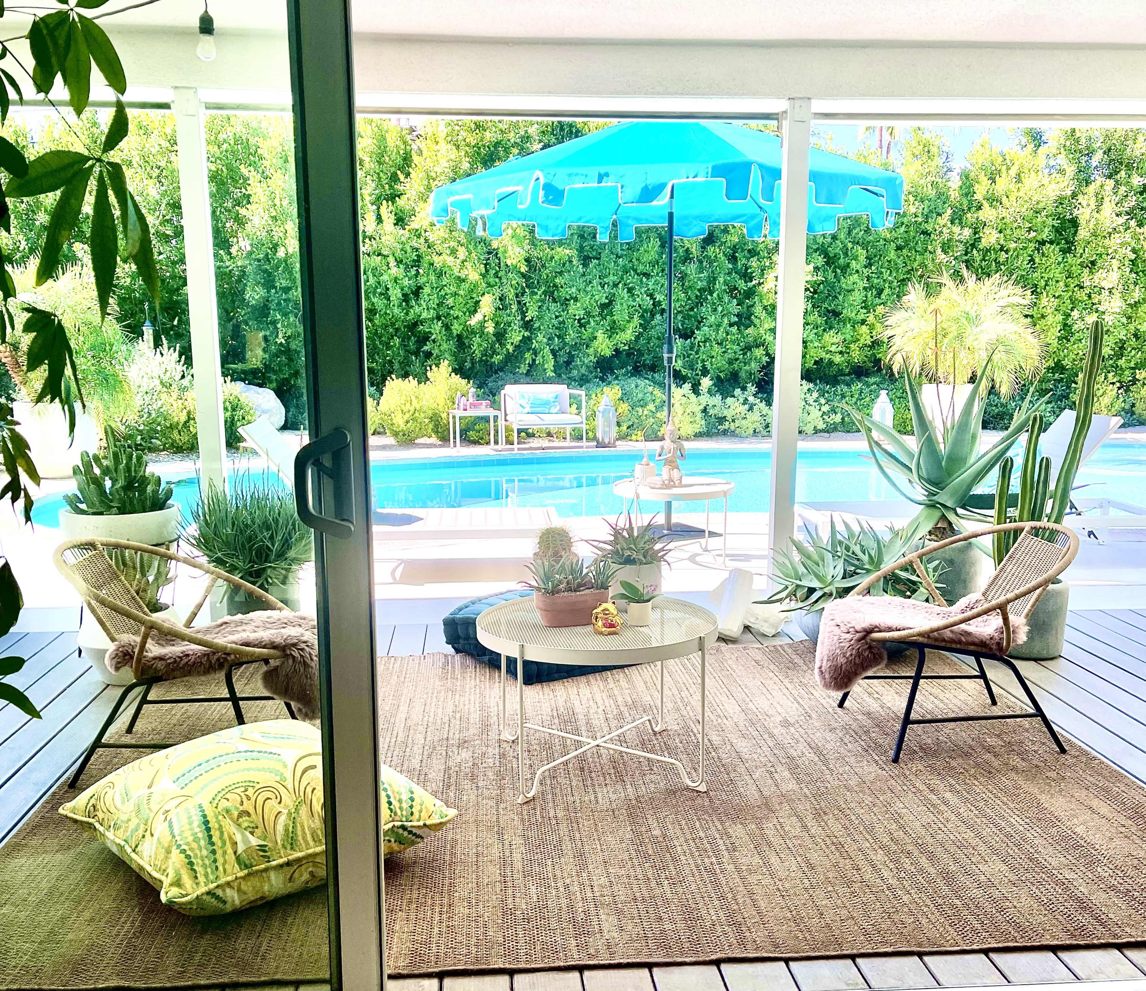 The scene features a covered outdoor seating area with two chairs, a small table, and various plants, overlooking a swimming pool surrounded by greenery.