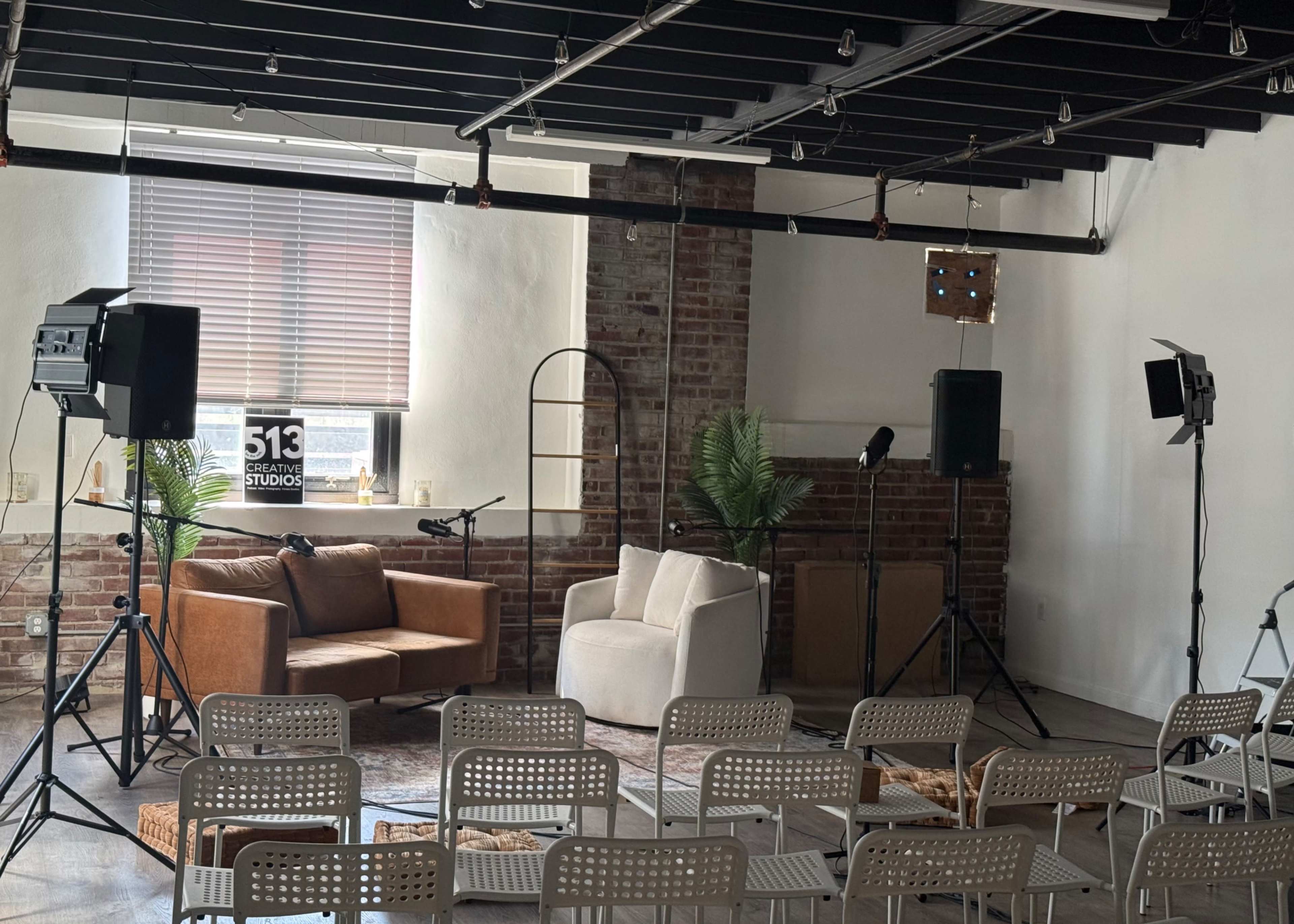 The image shows a small, staged event space with a sofa and a chair, surrounded by empty chairs for an audience, and equipped with audio equipment.