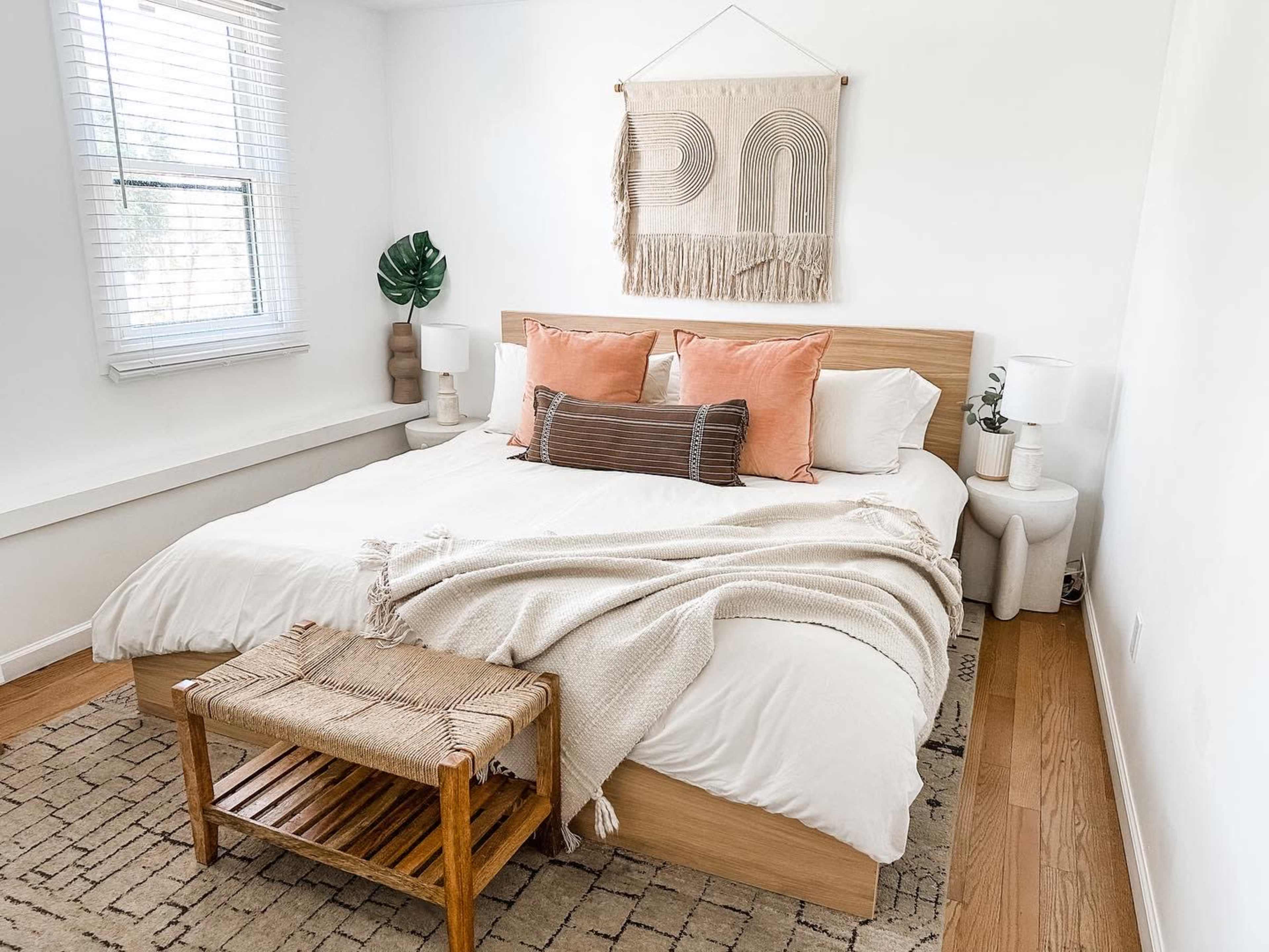The image depicts a neatly arranged bedroom featuring a wooden bed dressed with white linens and decorative pillows, accompanied by a woven blanket and a wooden side table.