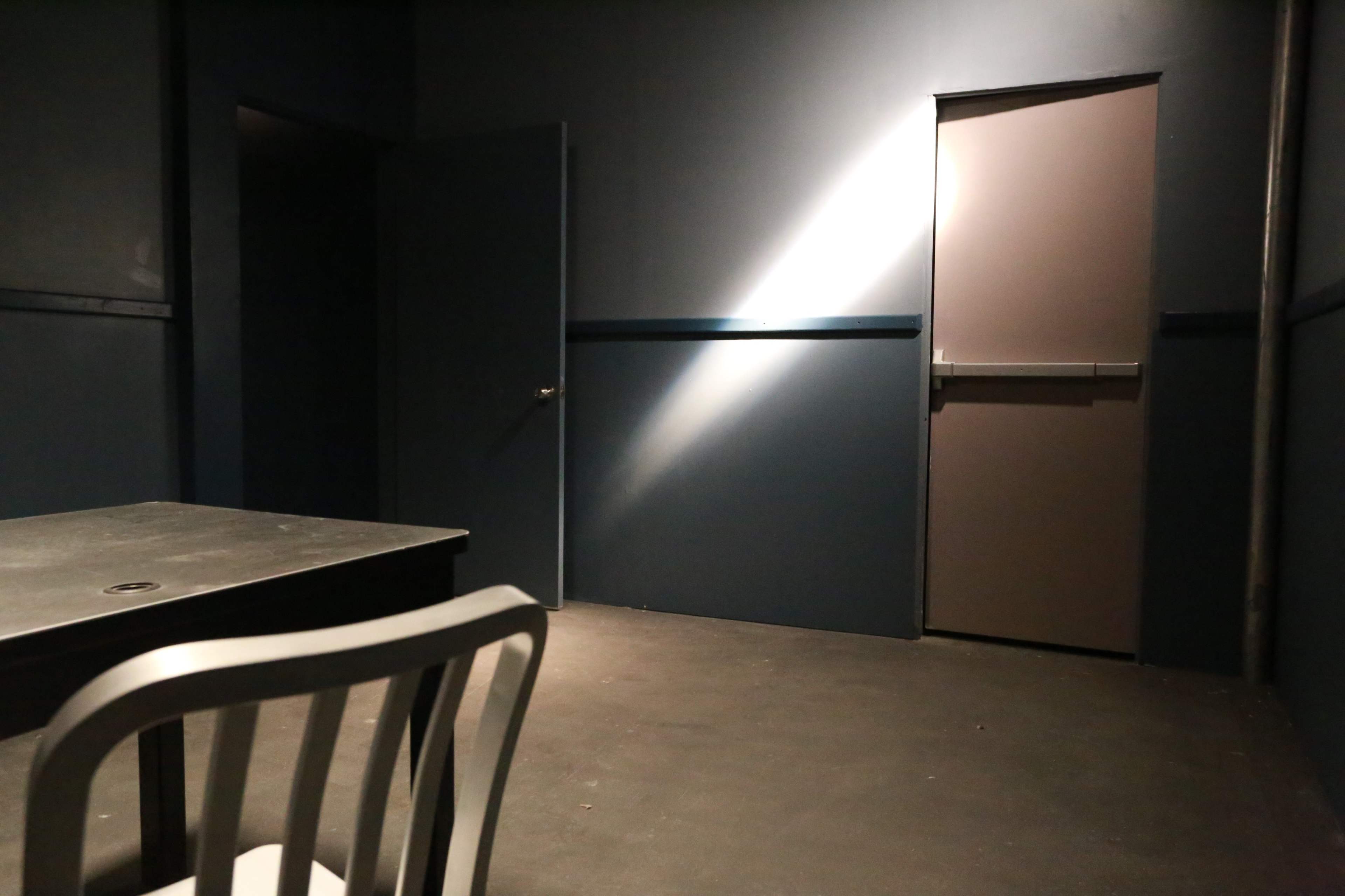 A dimly lit interrogation room features a table and a chair facing a closed door.