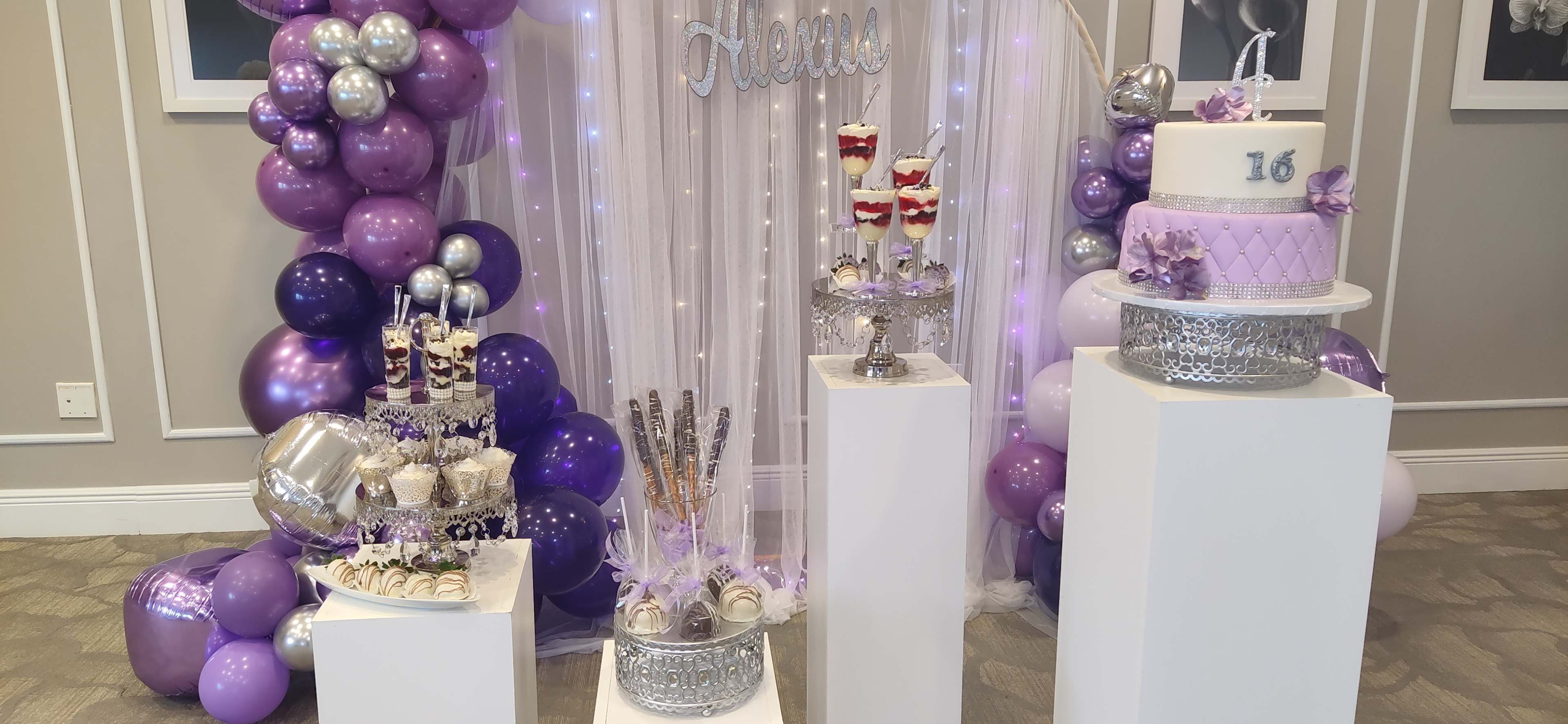 The image showcases a decorated event display featuring a large cake, dessert stands with sweet treats, and a backdrop of purple and silver balloons arranged in an elegant setting.