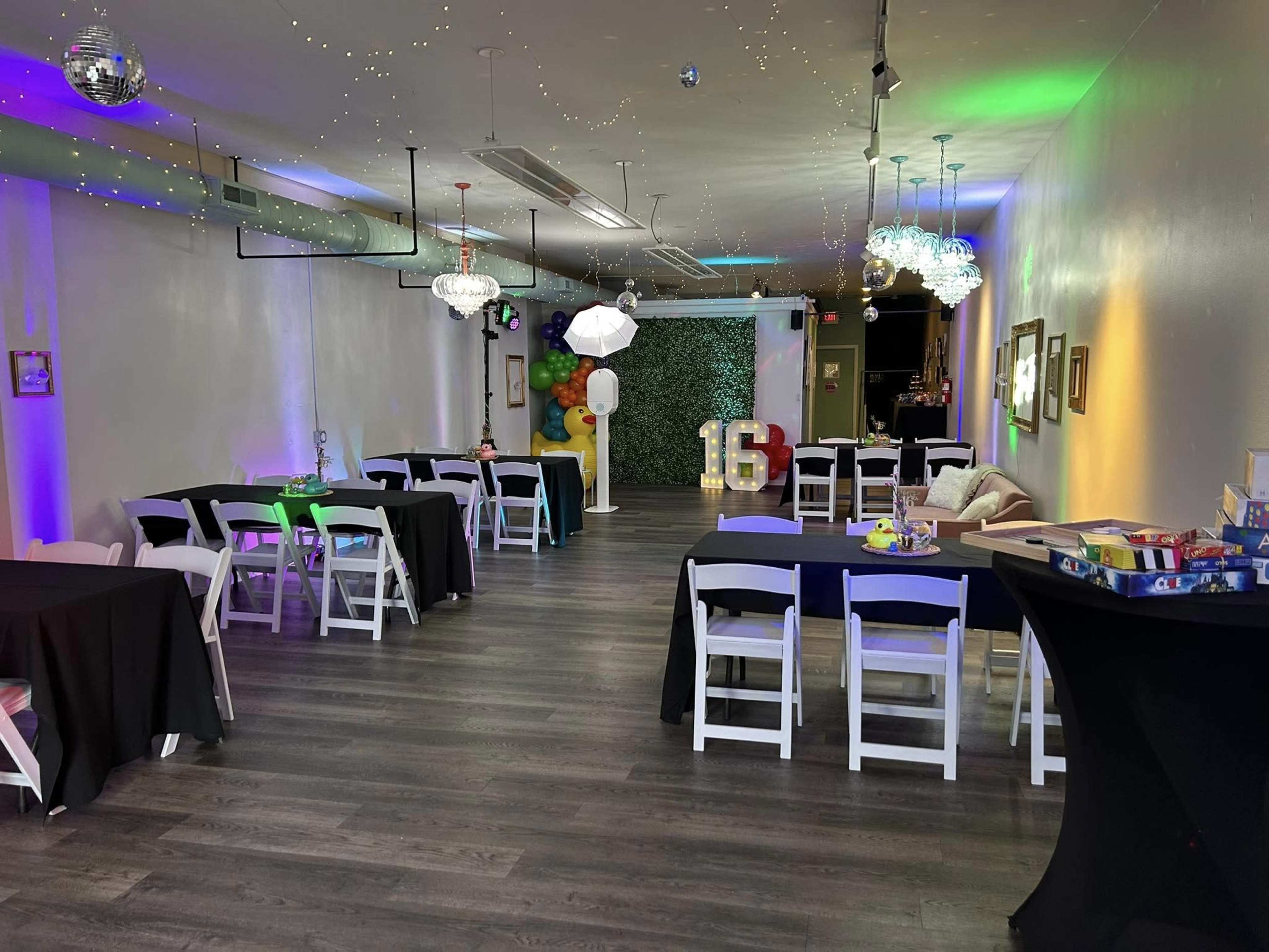 A spacious event room is set up with black and white tables, colorful decorations, and ambient lighting for a celebration.