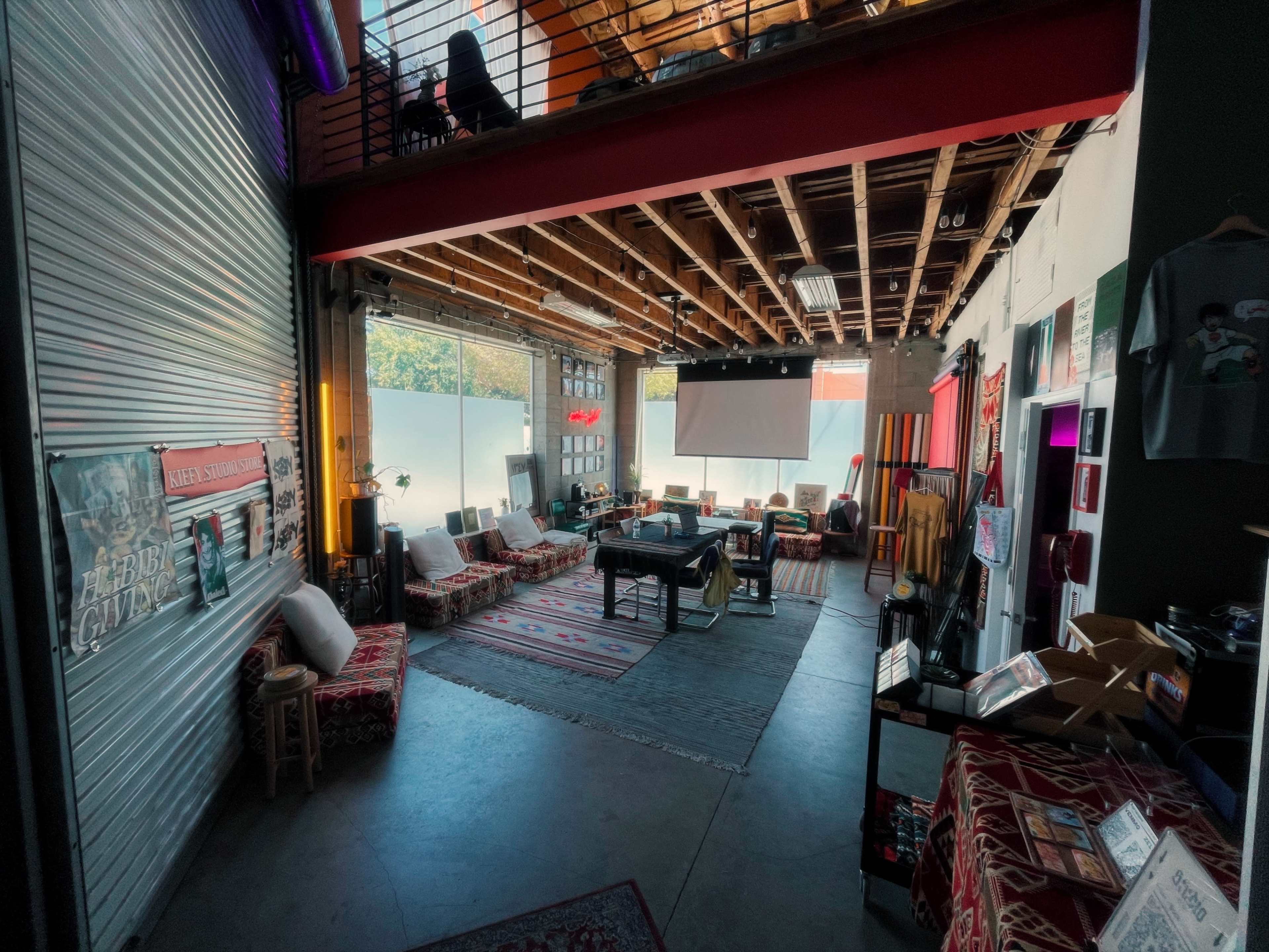 The image shows a spacious, contemporary room with a mix of seating arrangements, decorative rugs, and a large screen at one end.