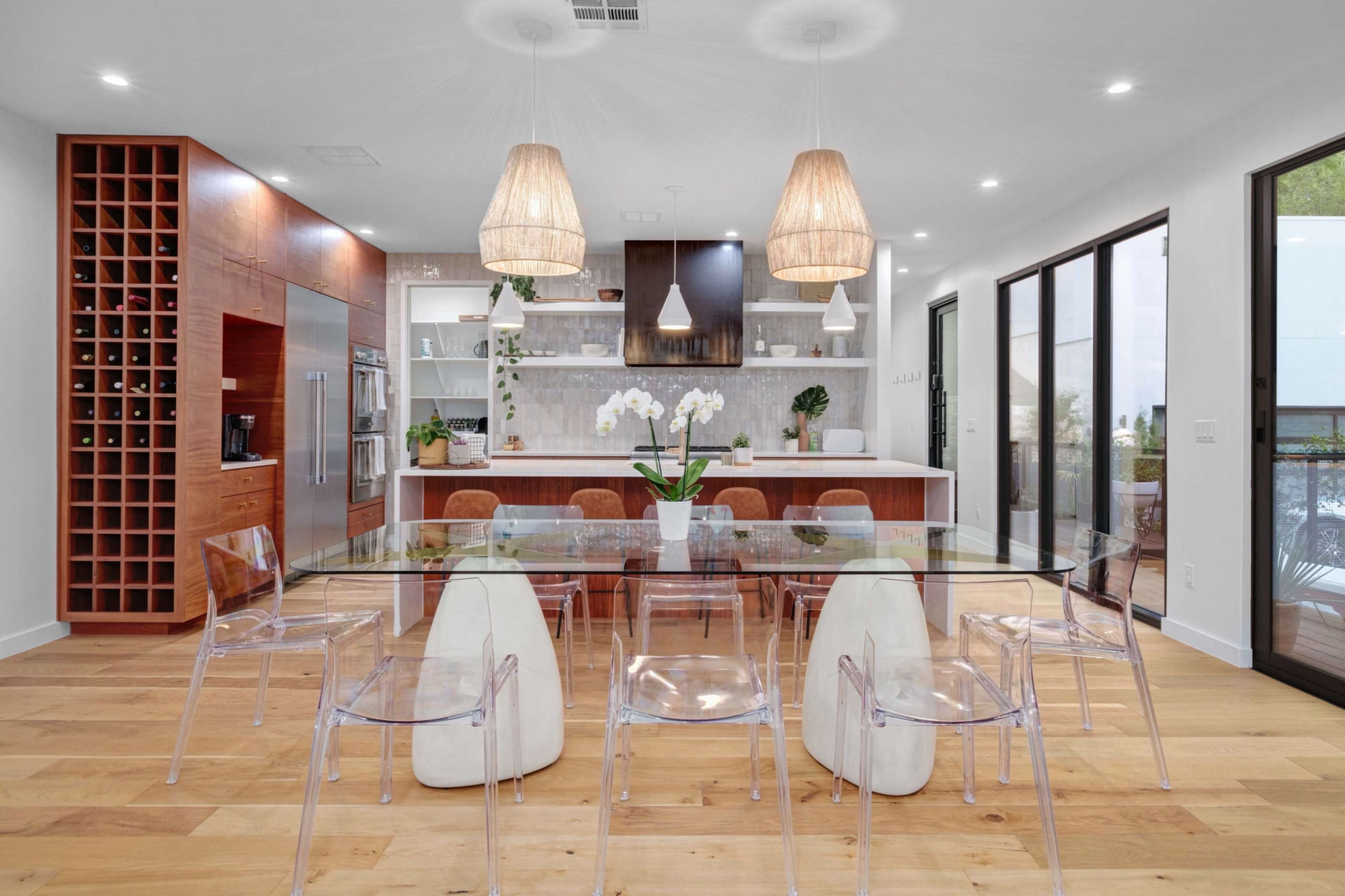 A modern kitchen and dining area with a glass dining table surrounded by clear acrylic chairs, wooden cabinetry, and pendant lighting.