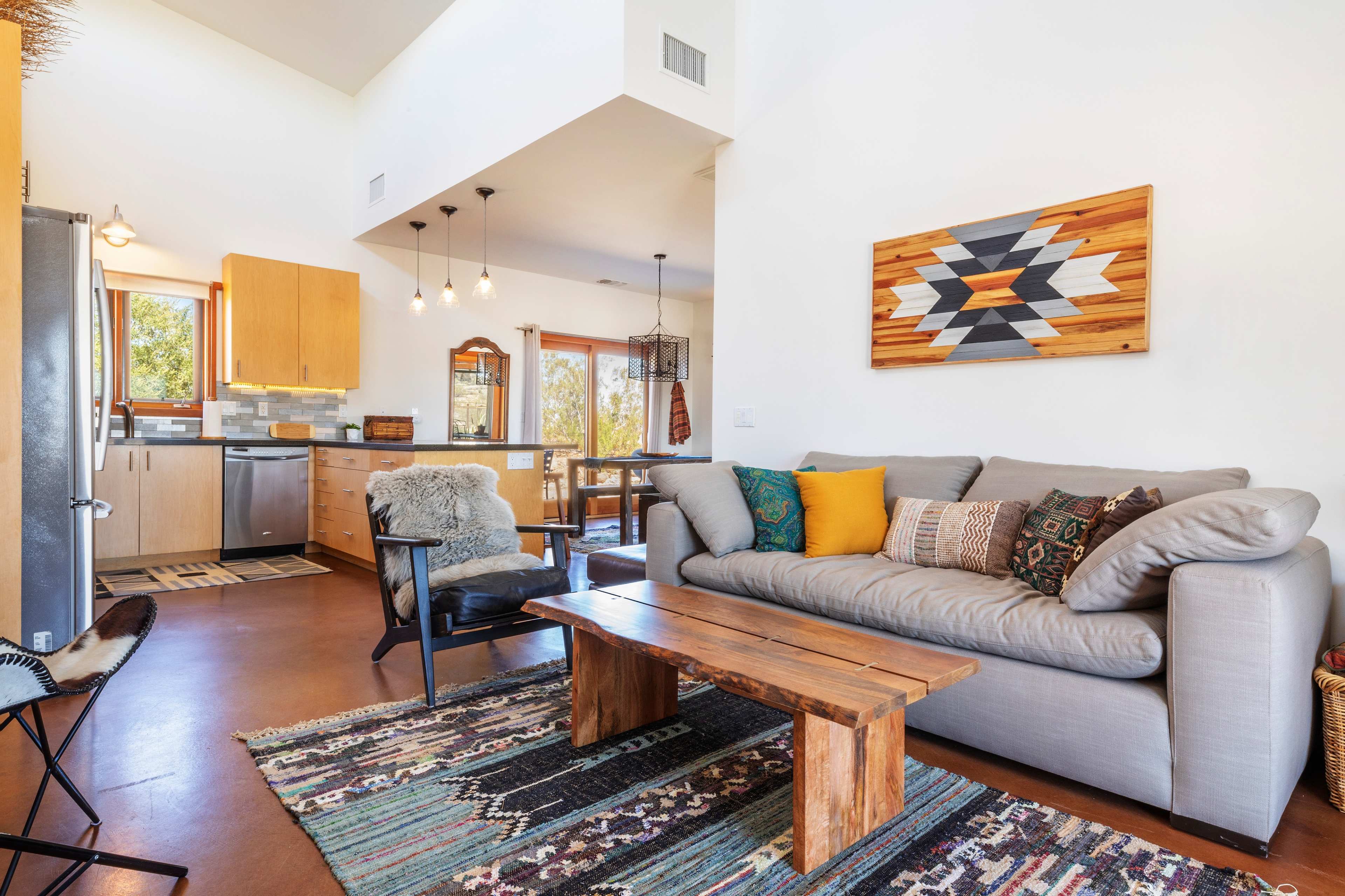 A modern living room features a gray sofa with colorful cushions, a wooden coffee table, and artwork on the wall, alongside an open kitchen with wooden cabinets and large windows.