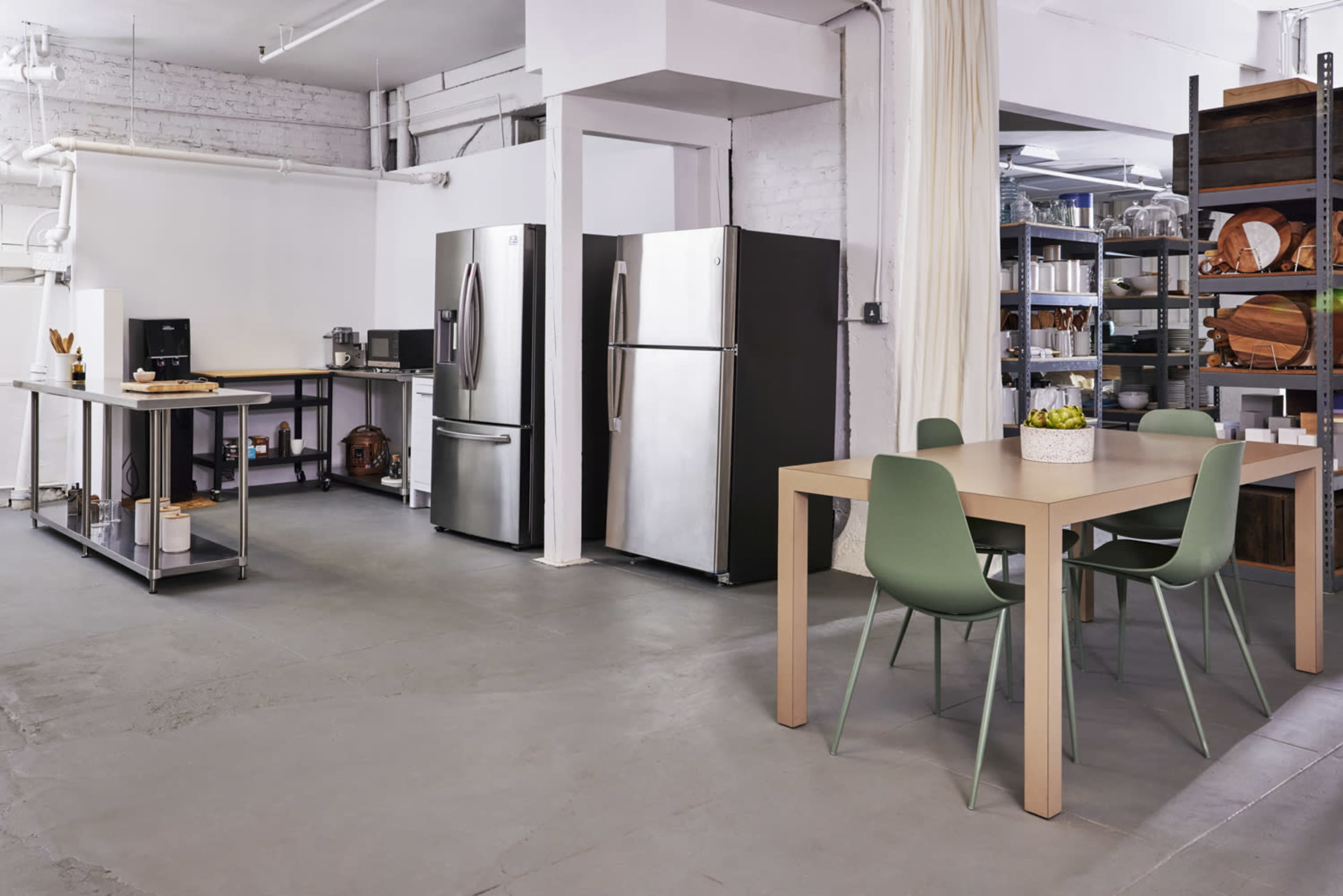 A modern kitchen space featuring stainless steel refrigerators, a dining table with green chairs, and a shelving unit filled with various kitchen items.