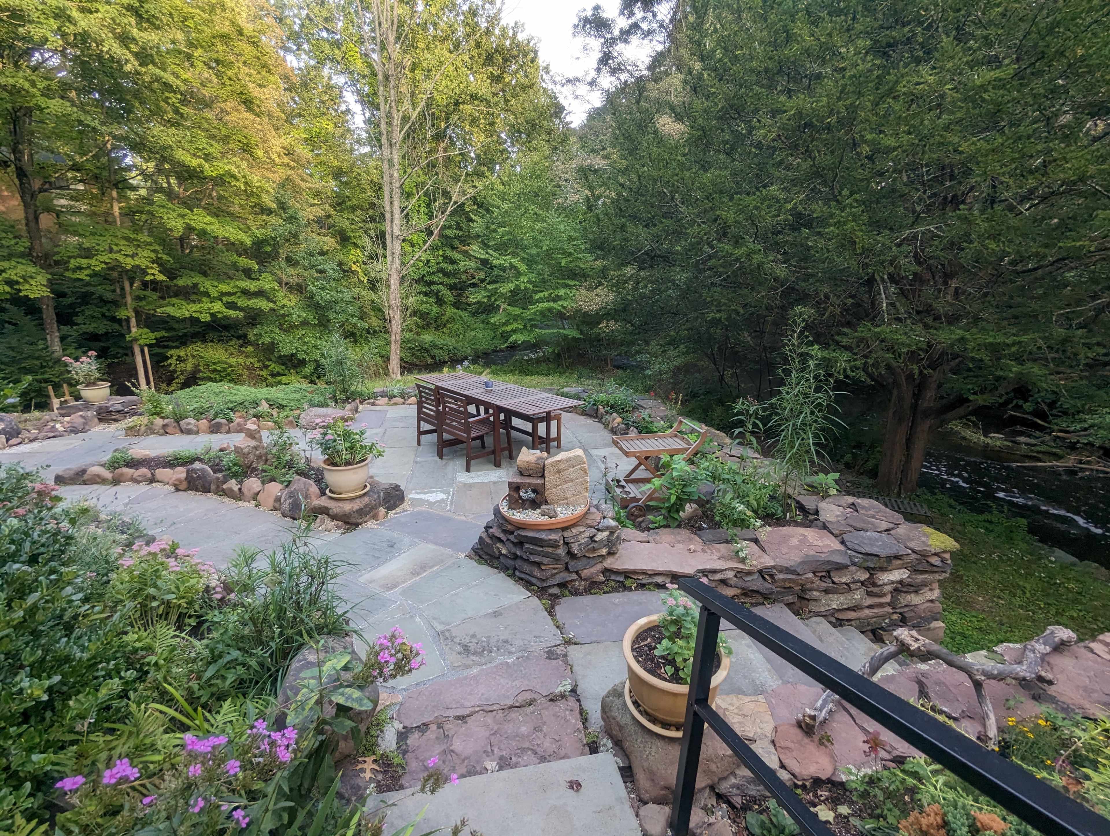 A landscaped outdoor area with a stone patio, a wooden table and chairs, and surrounding greenery beside a stream.