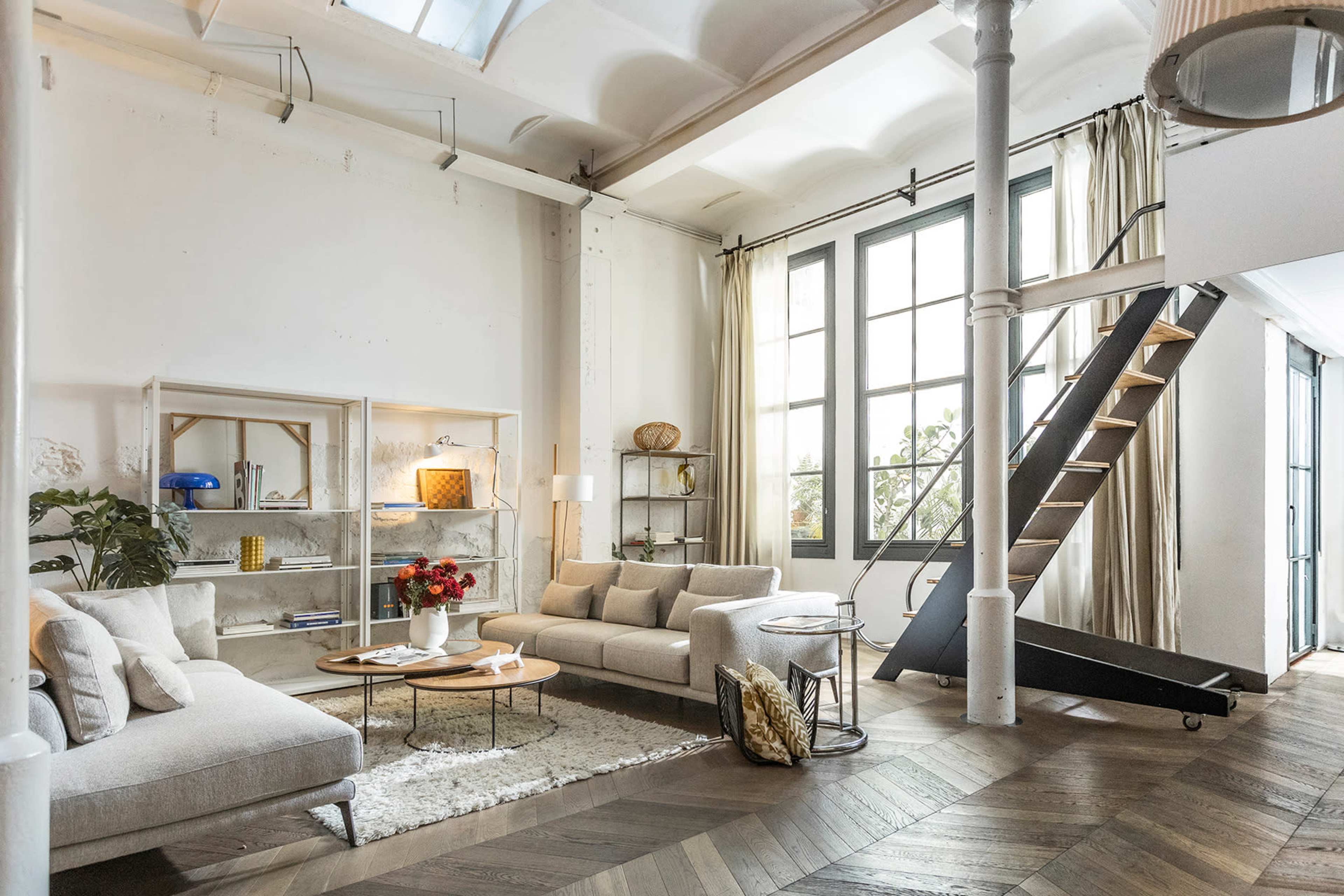 The image shows a modern living room with a light-colored sofa, a coffee table, and a staircase leading to a loft area beside large windows.