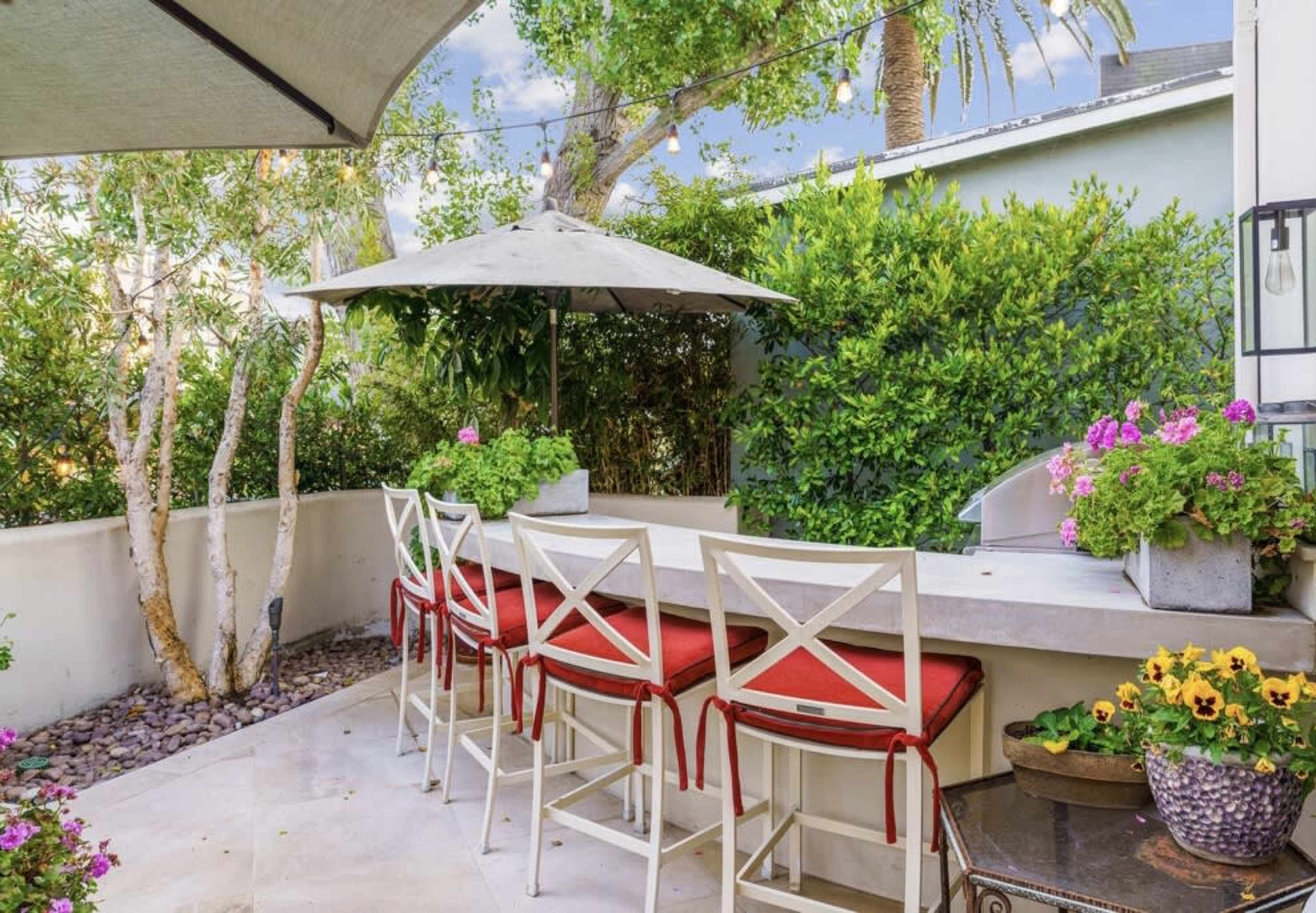 A small outdoor seating area features a bar with four white stools and an umbrella, surrounded by lush greenery and colorful flowers.
