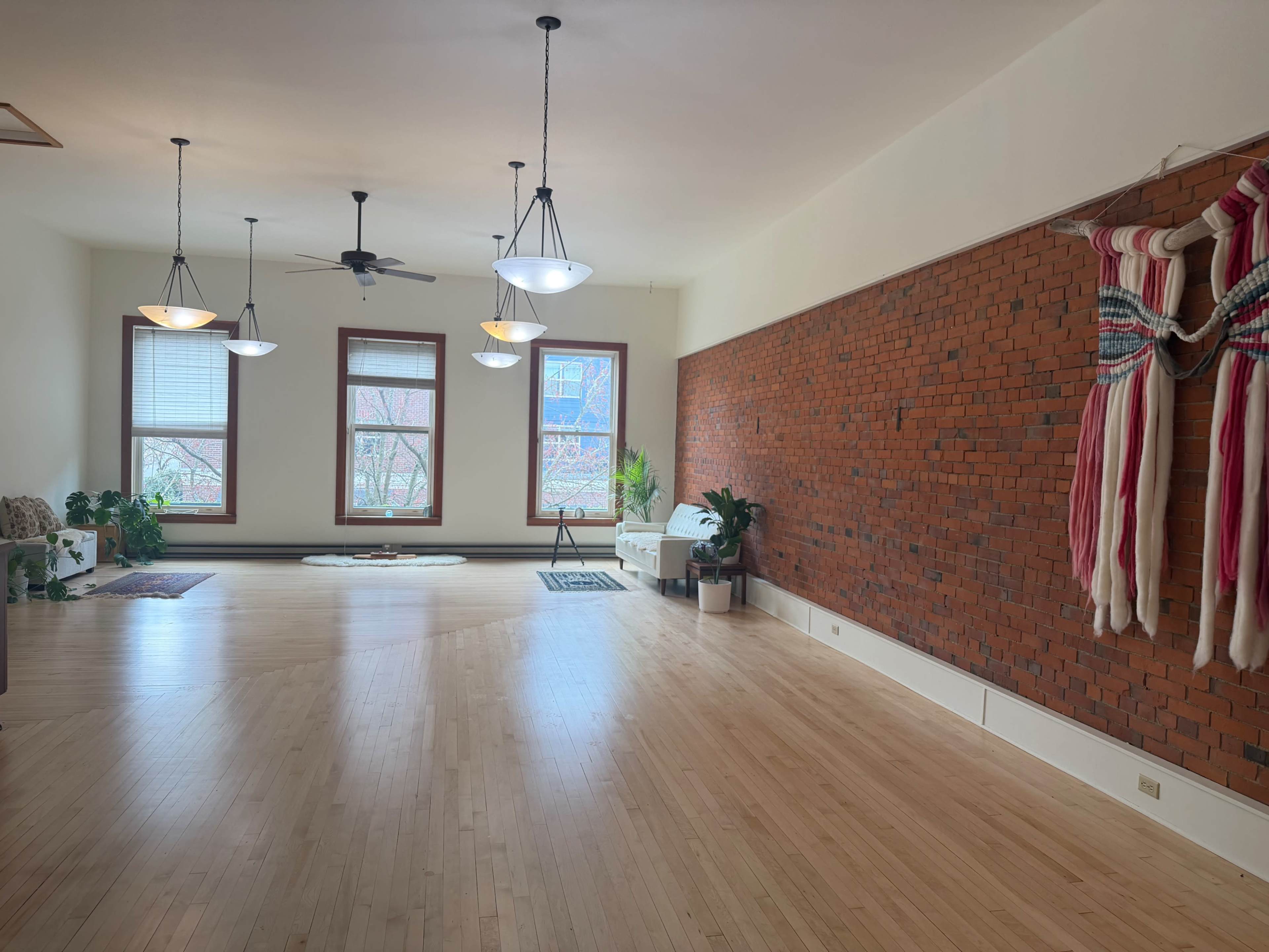 The image shows a spacious, well-lit room with hardwood floors, exposed brick walls, and large windows, featuring ceiling fans and decorative wall hangings.