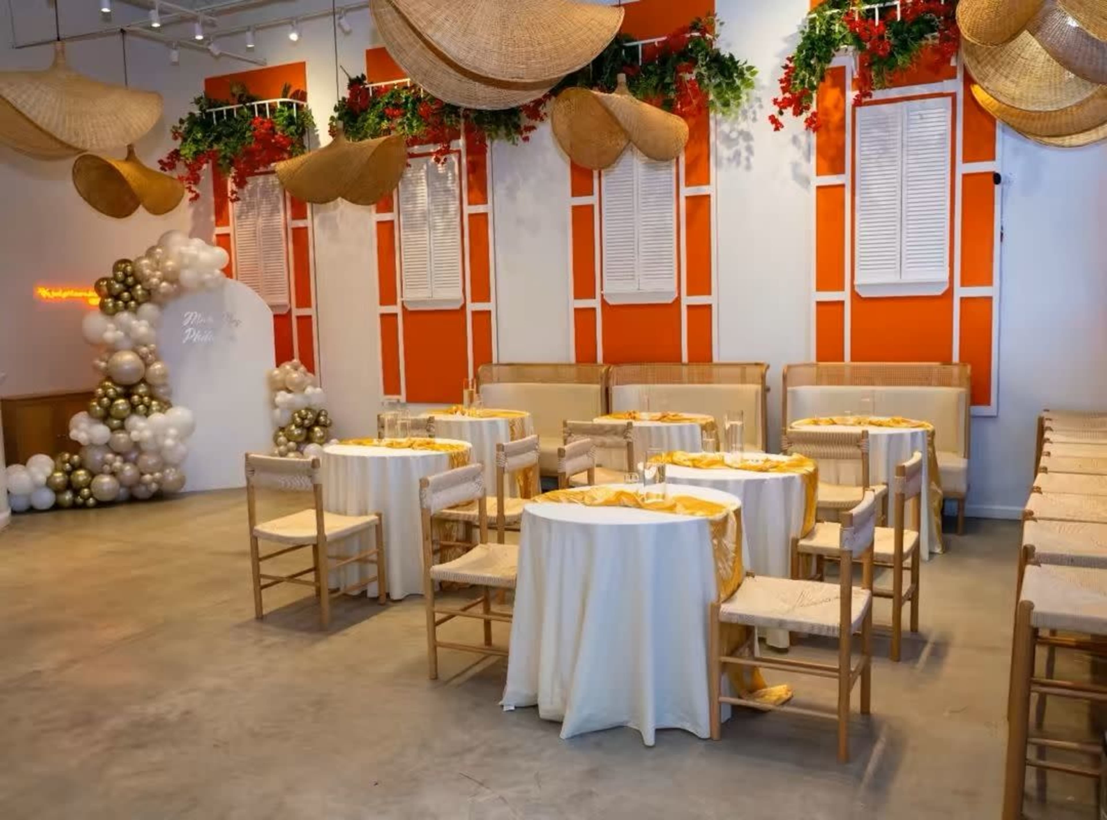 The image depicts a spacious event venue with decorated tables covered in white tablecloths and golden accents, surrounded by colorful wall decorations and greenery.