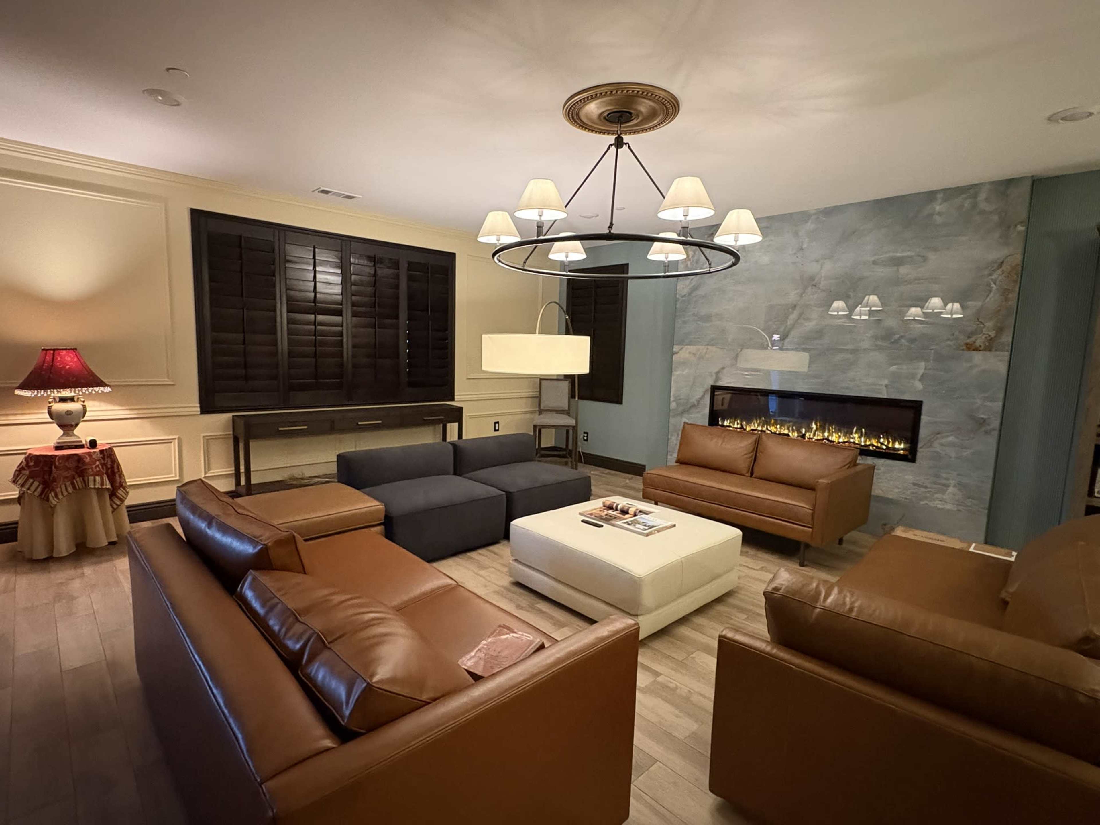 The image shows a modern living room with brown leather sofas arranged around a central coffee table, facing a wall-mounted fireplace, and featuring warm lighting fixtures.