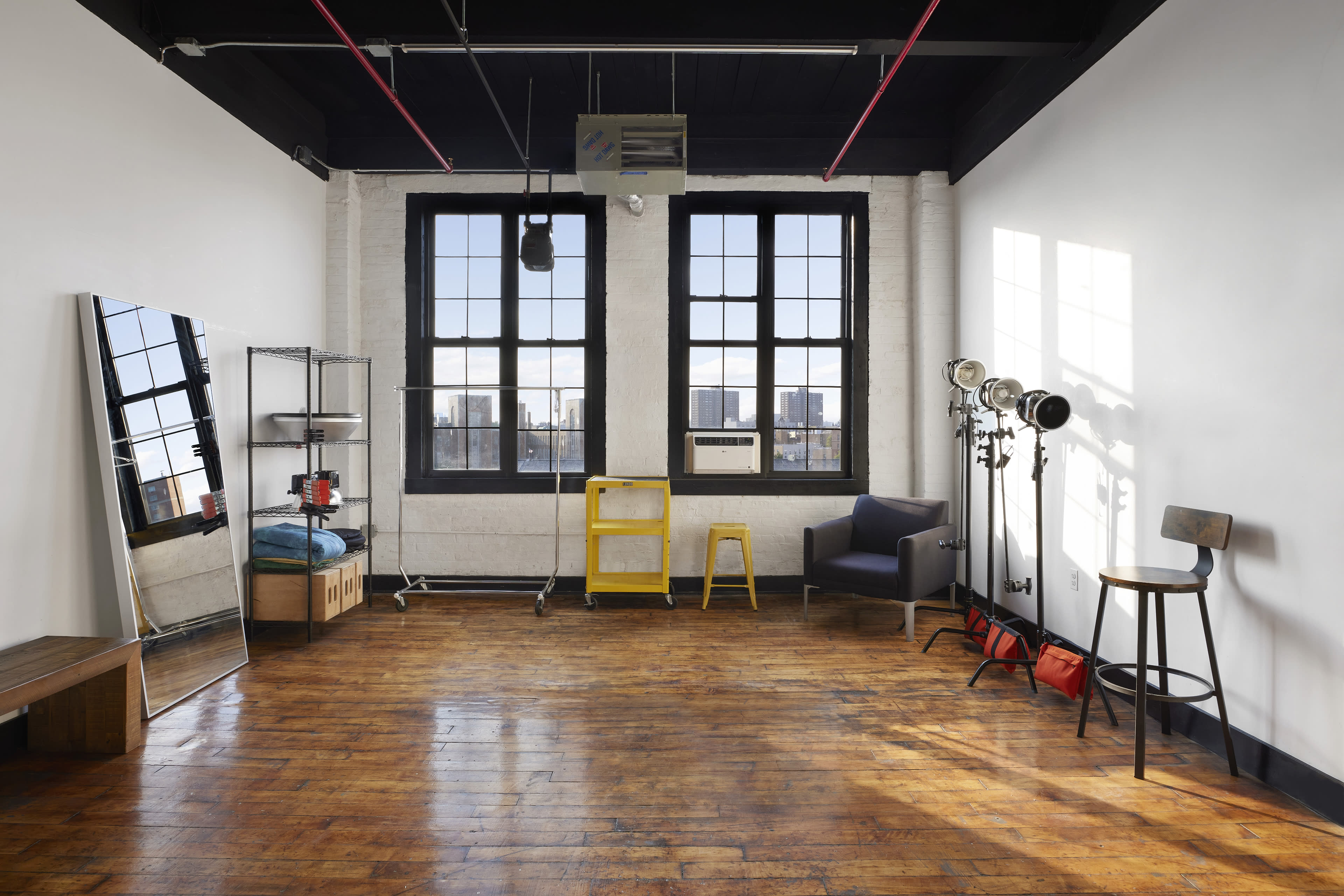 A spacious room features large windows, wooden flooring, a mirror, various seating options, and lighting equipment.