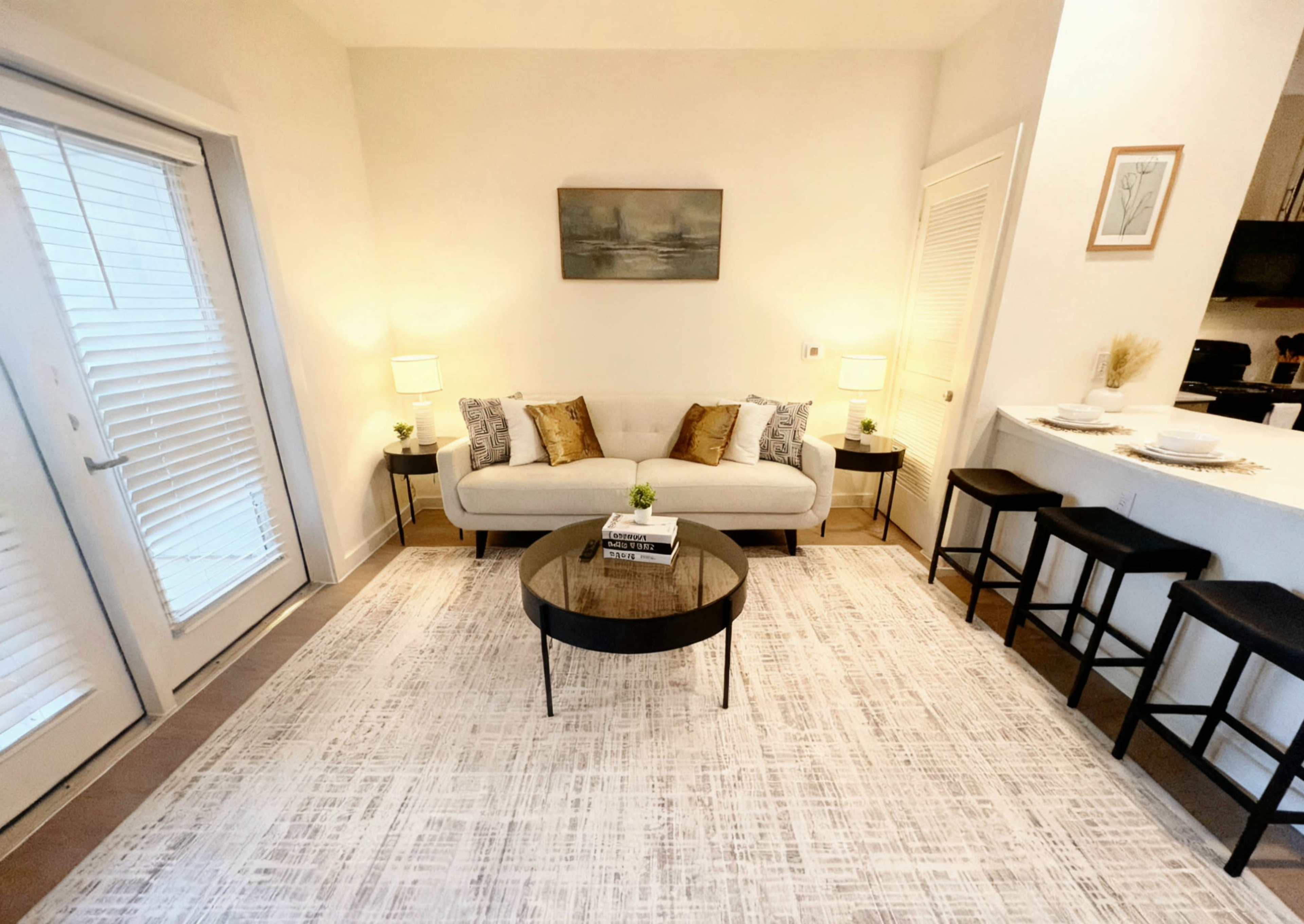 The image shows a cozy living area with a light-colored couch, a round coffee table, and two side tables with lamps, all arranged around a modern rug and facing an open kitchen space.