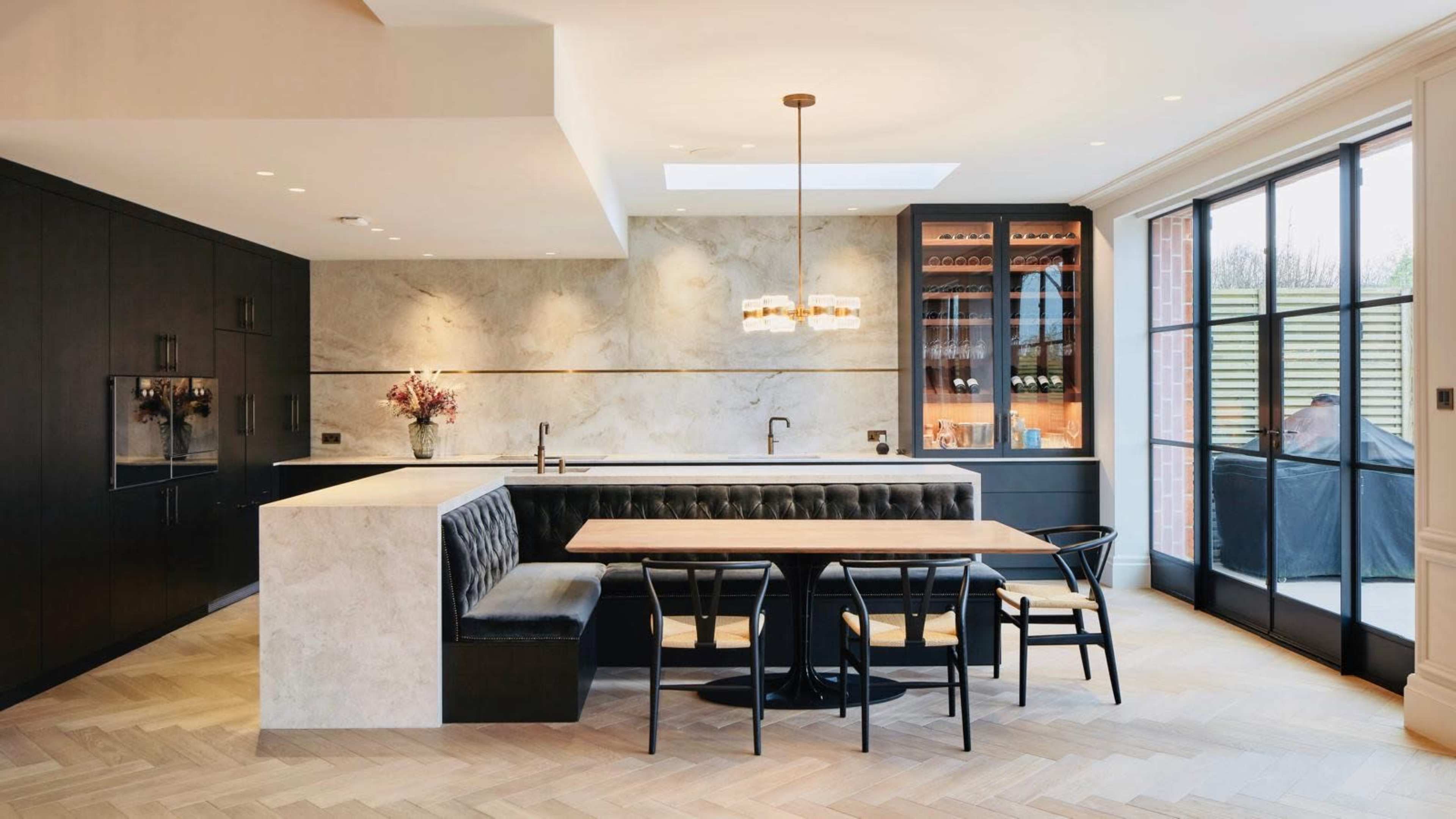 The image depicts a modern kitchen with marble countertops, a black leather banquette, a wooden dining table, and large windows allowing natural light to enter the space.