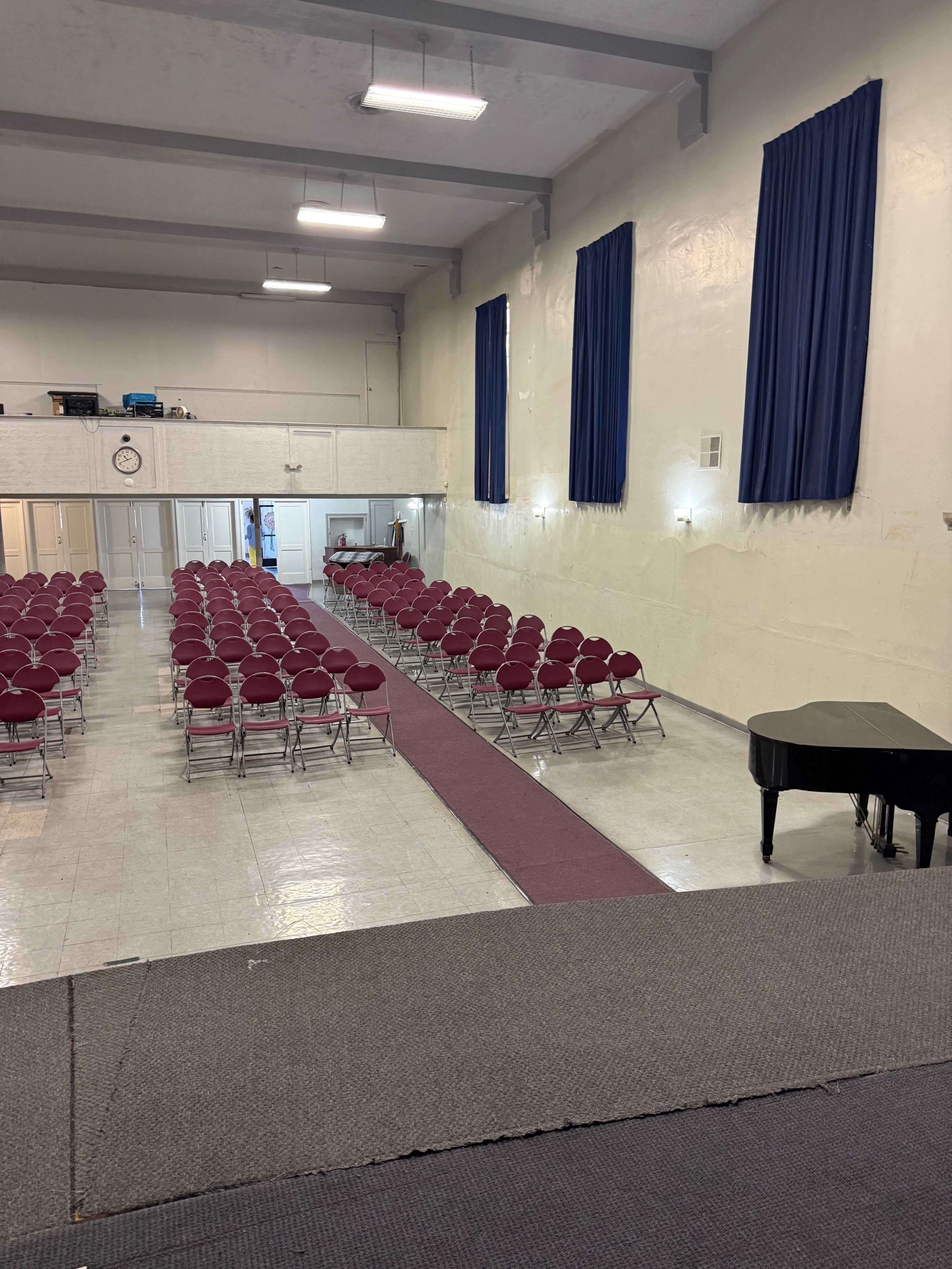 The image shows a large room with rows of maroon chairs facing a stage, separated by a red carpet, and flanked by blue curtains on the walls.