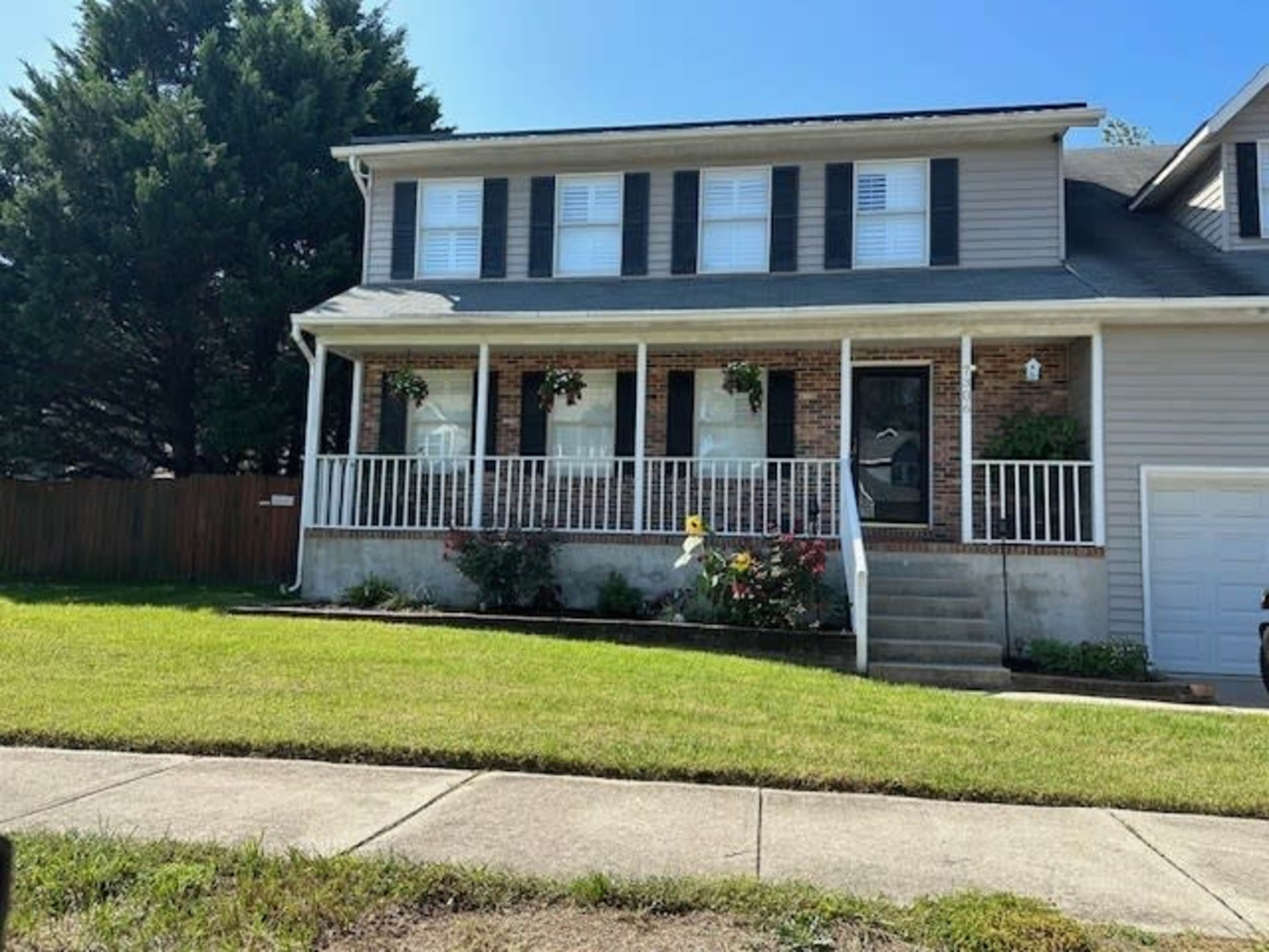 The image shows a two-story house with a front porch, several flower pots hanging from the railing, and a neatly trimmed lawn.