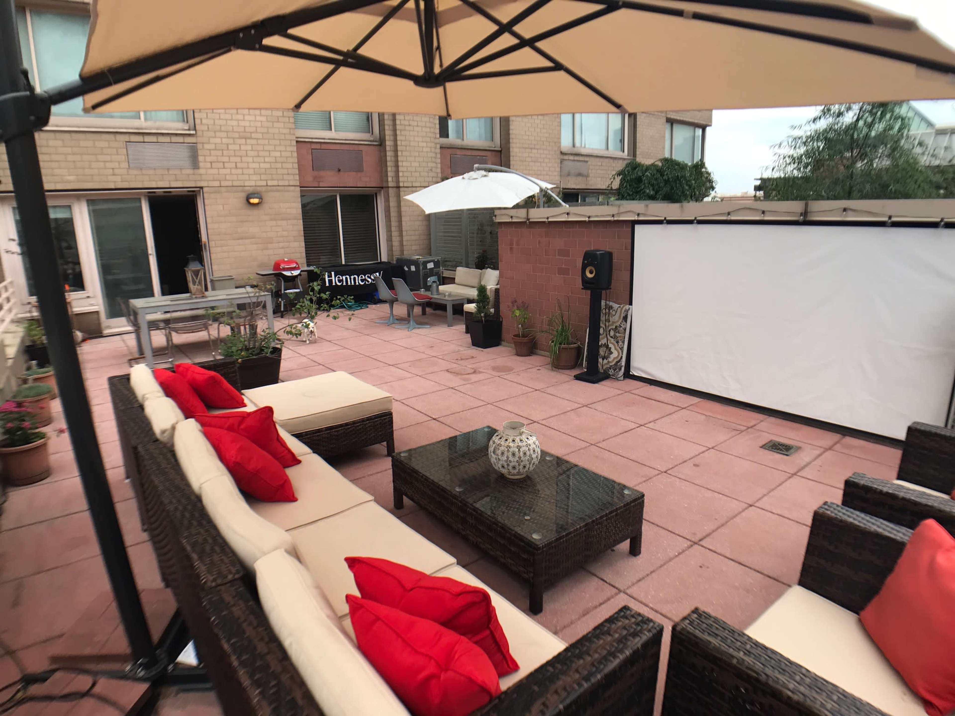 A patio area with a sectional sofa, coffee table, and a large white screen for projected viewing, surrounded by potted plants and an outdoor umbrella.