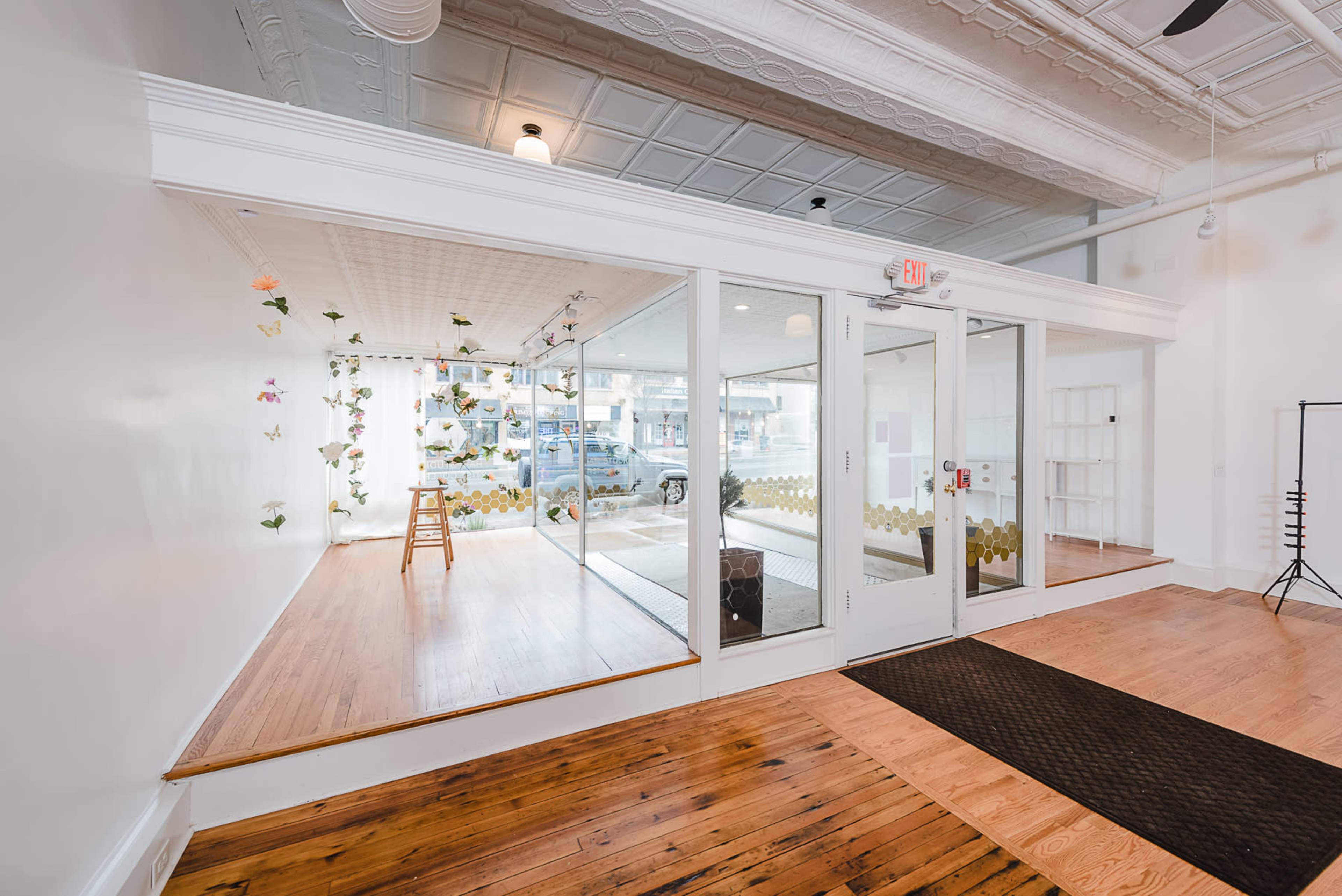 A bright, modern space features large glass windows, wooden flooring, and decorative elements like plants and a stool inside.