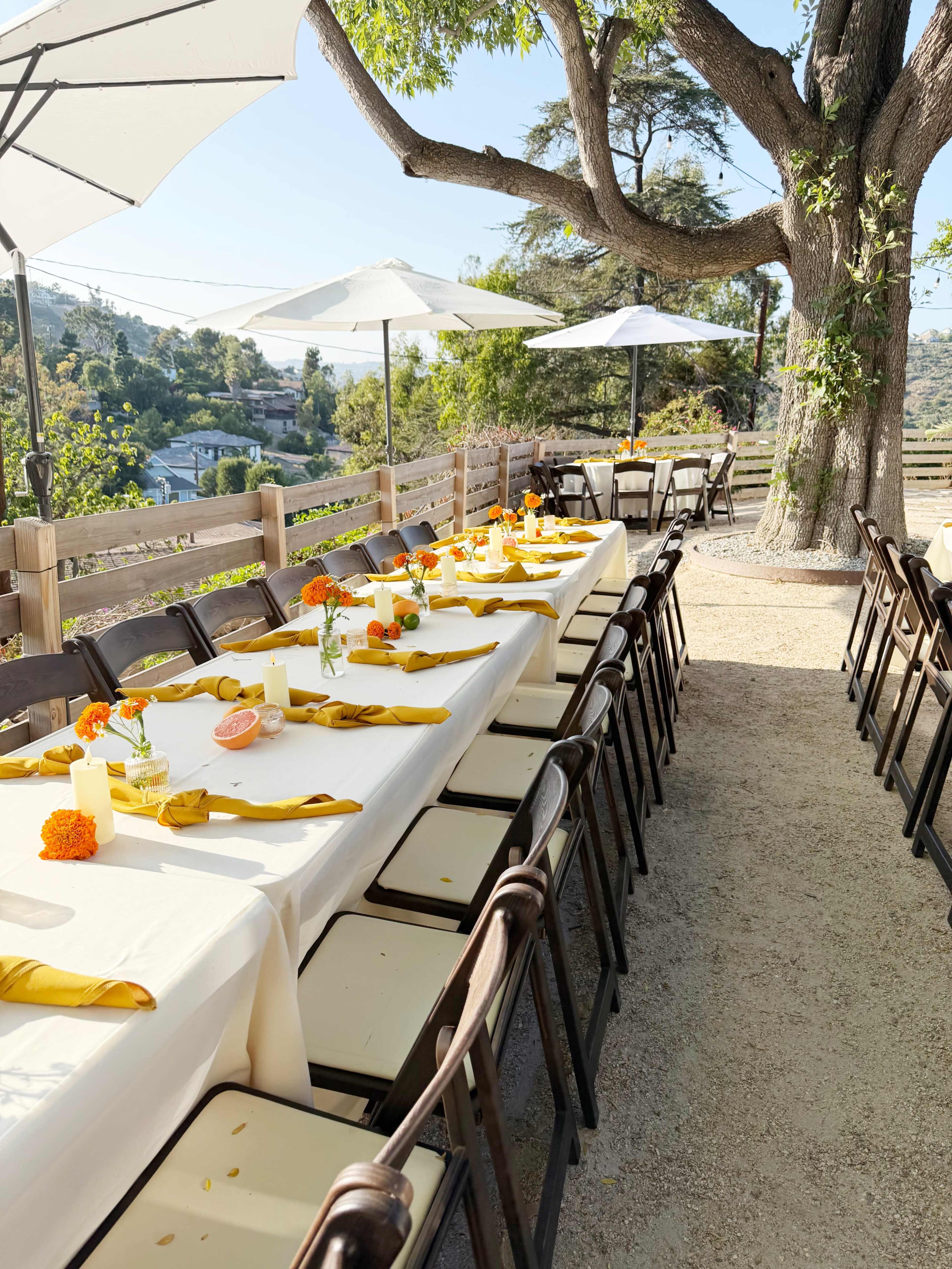 A long table set for an outdoor event is arranged under large umbrellas, with orange flowers and yellow napkins placed neatly at each setting.
