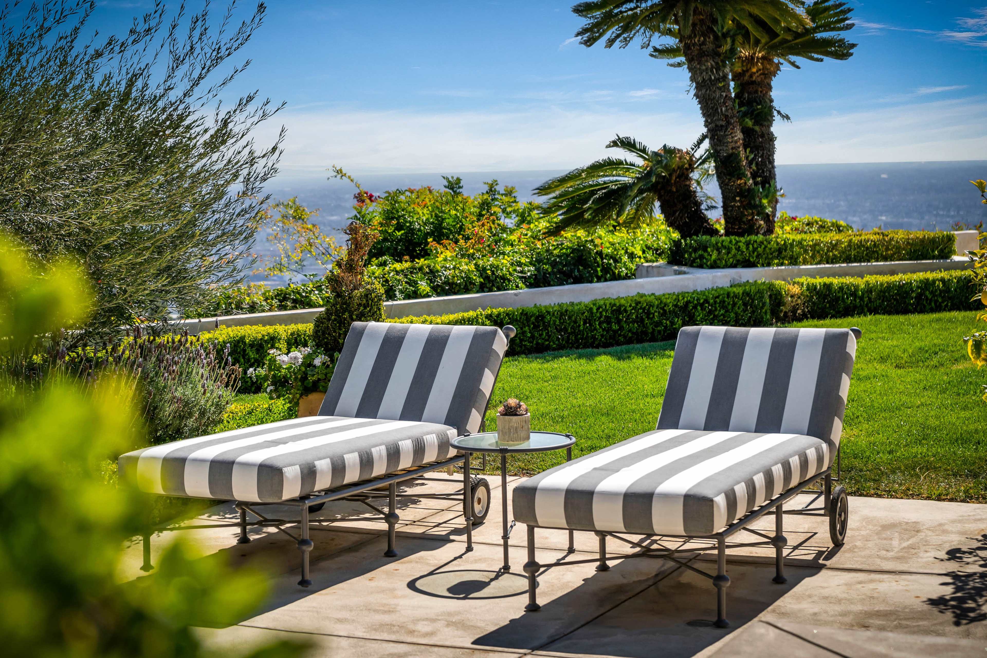 Two striped lounge chairs are positioned beside a small table against a backdrop of lush greenery and a clear blue sky.