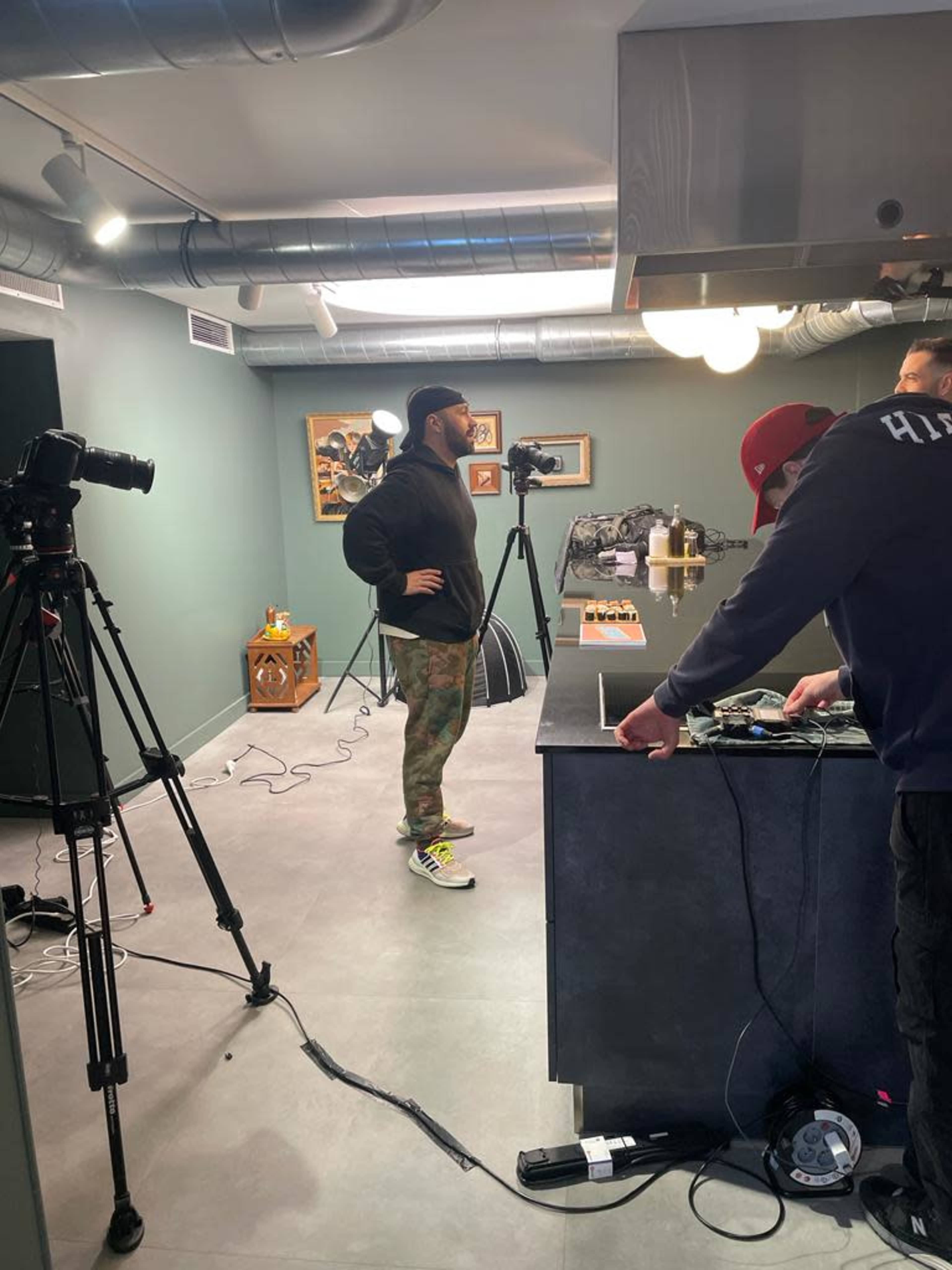 A man stands with his hands on his hips in a studio kitchen, while two cameras are set up nearby and another person is working at the counter.