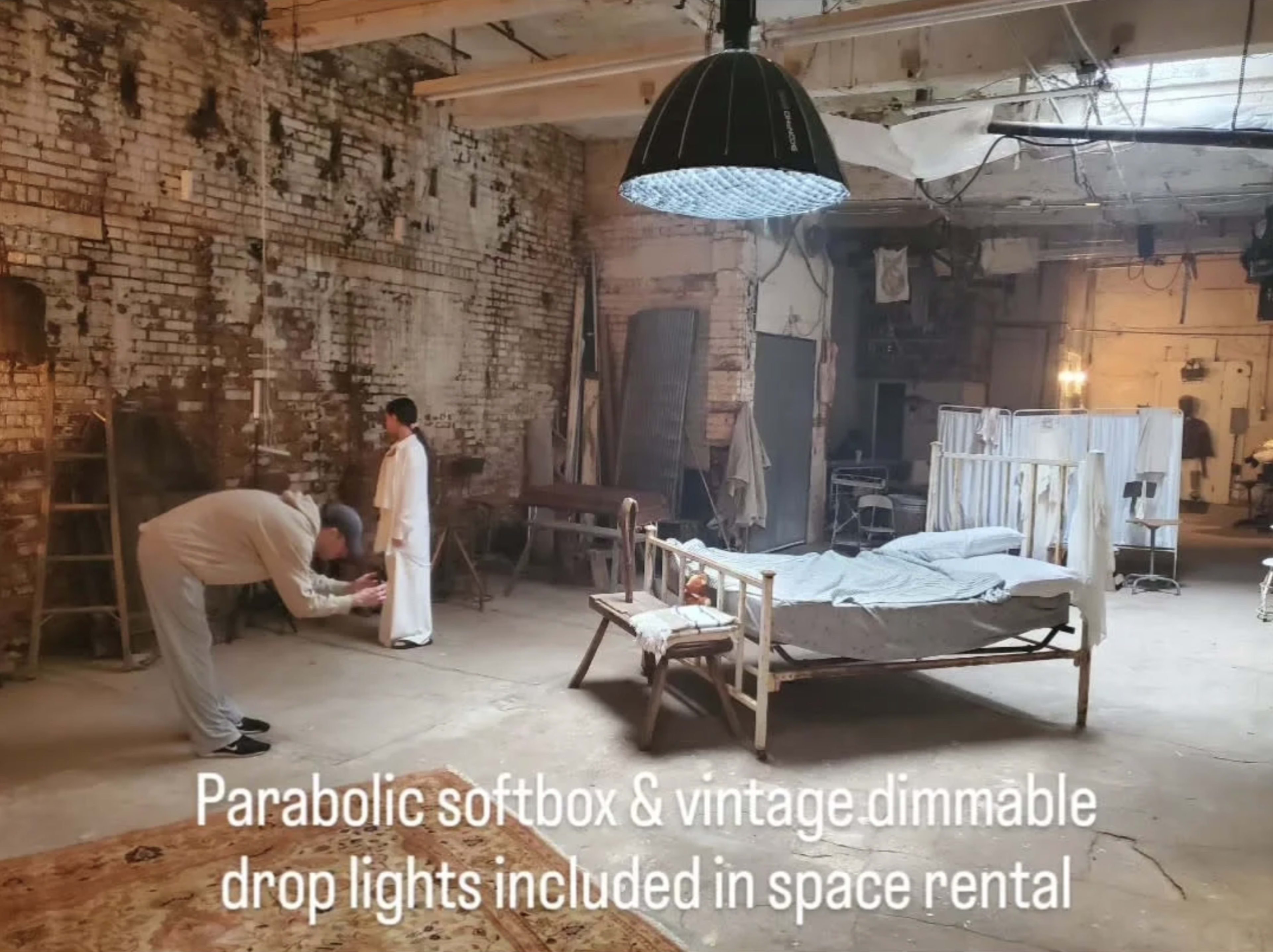 A spacious, industrial-style room features a bed, vintage furniture, and a person setting up lighting equipment.