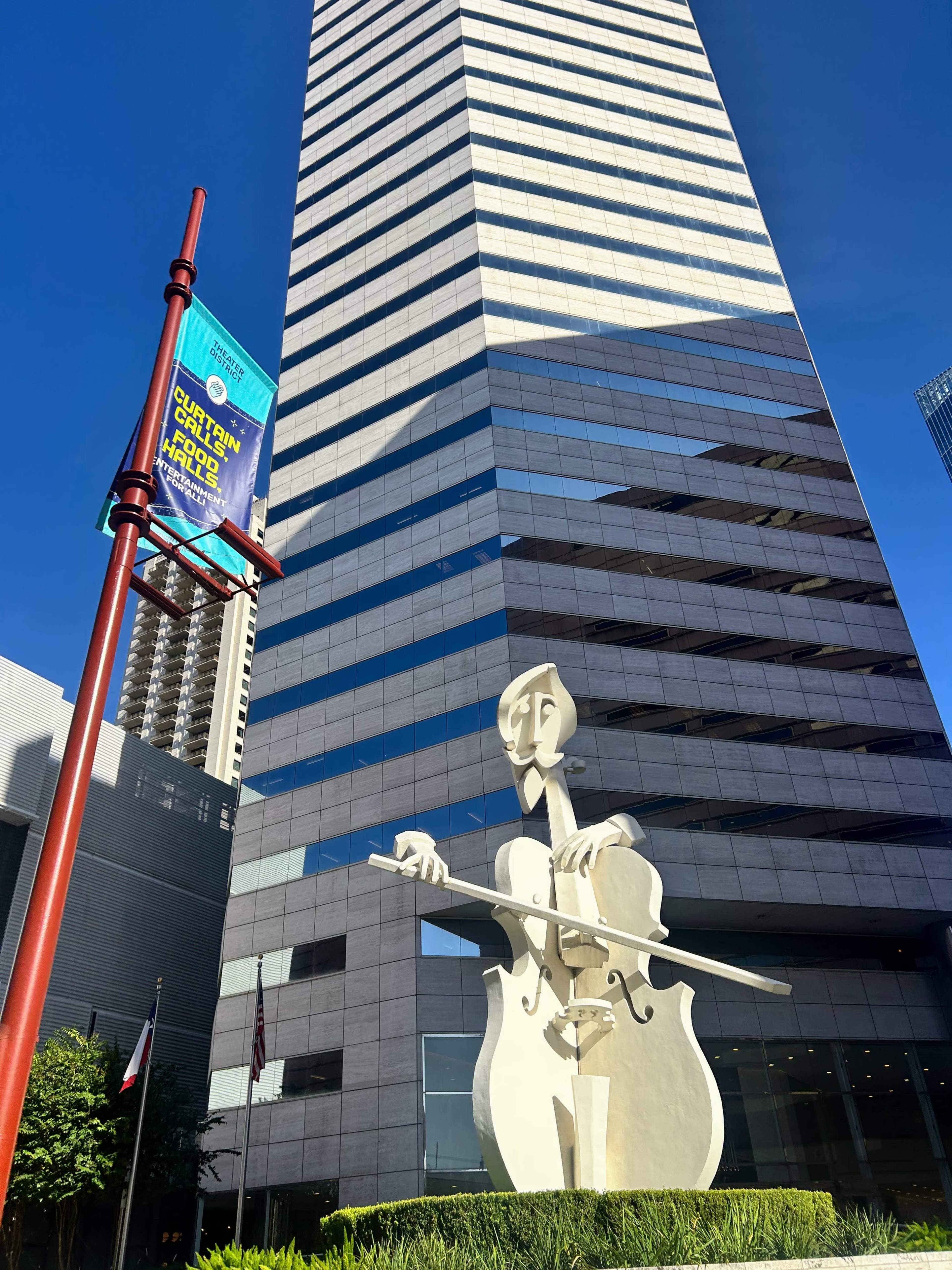A large white sculpture of a cellist stands in front of a tall, modern skyscraper, with a colorful banner nearby.