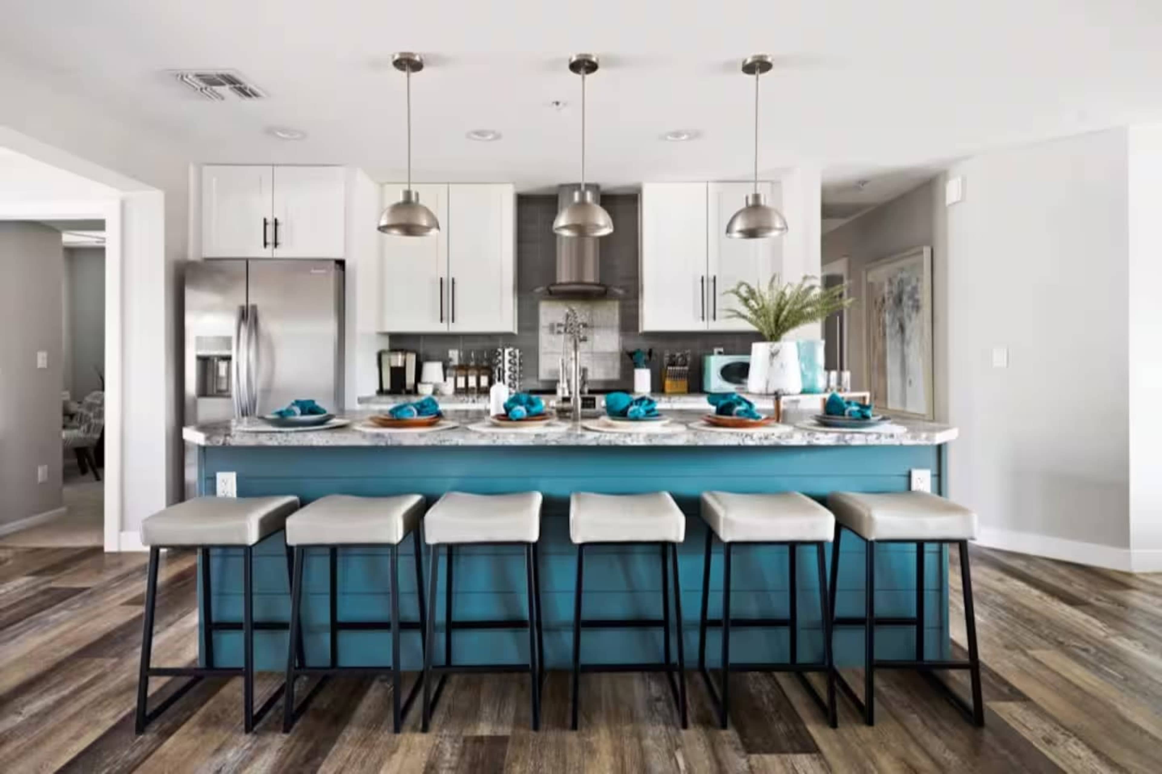 The kitchen features a central island with a blue base, accompanied by six beige bar stools and modern light fixtures overhead.