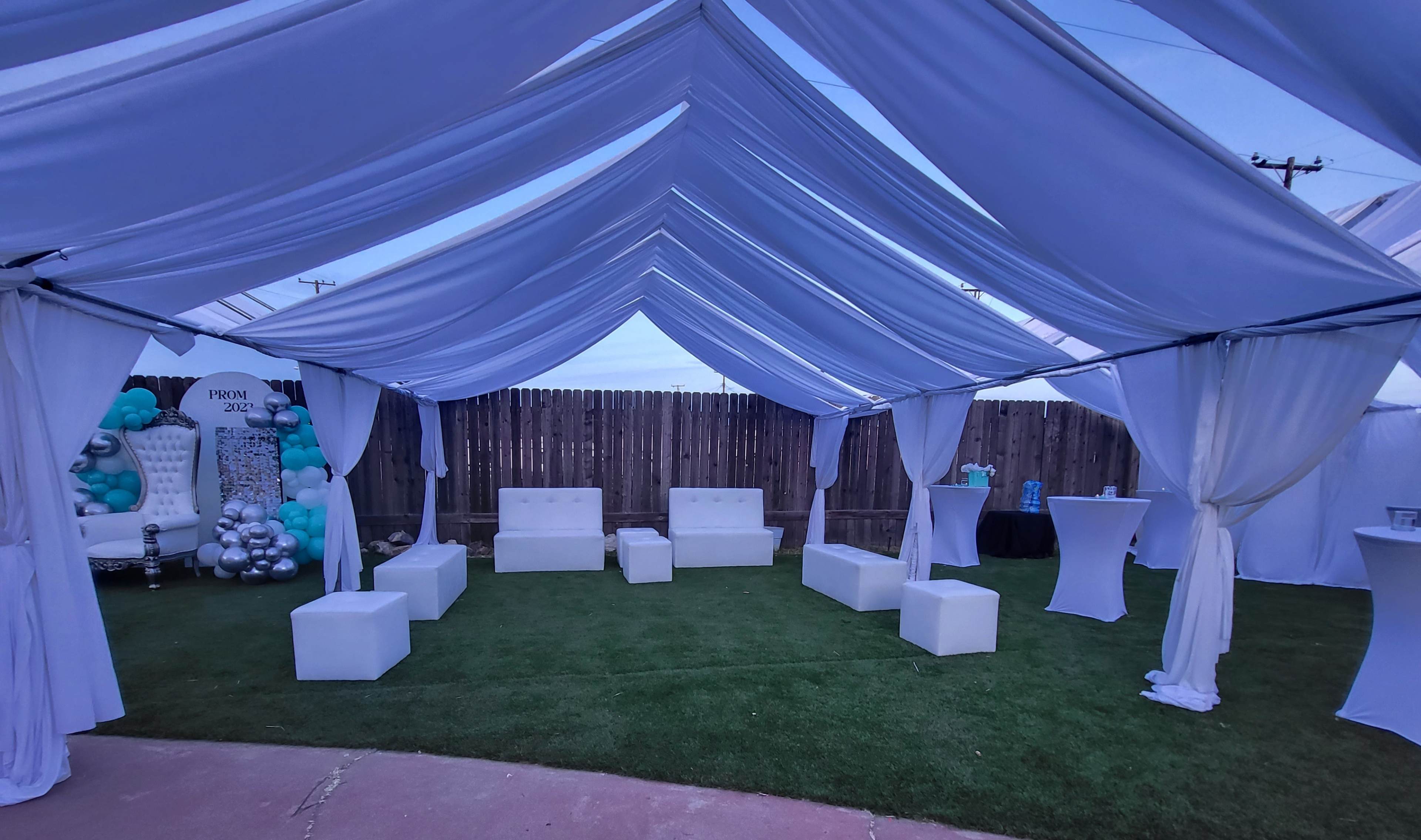 A beautifully arranged outdoor event space features white draped canopies, green grass, and white lounge seating with decorative elements in the background.