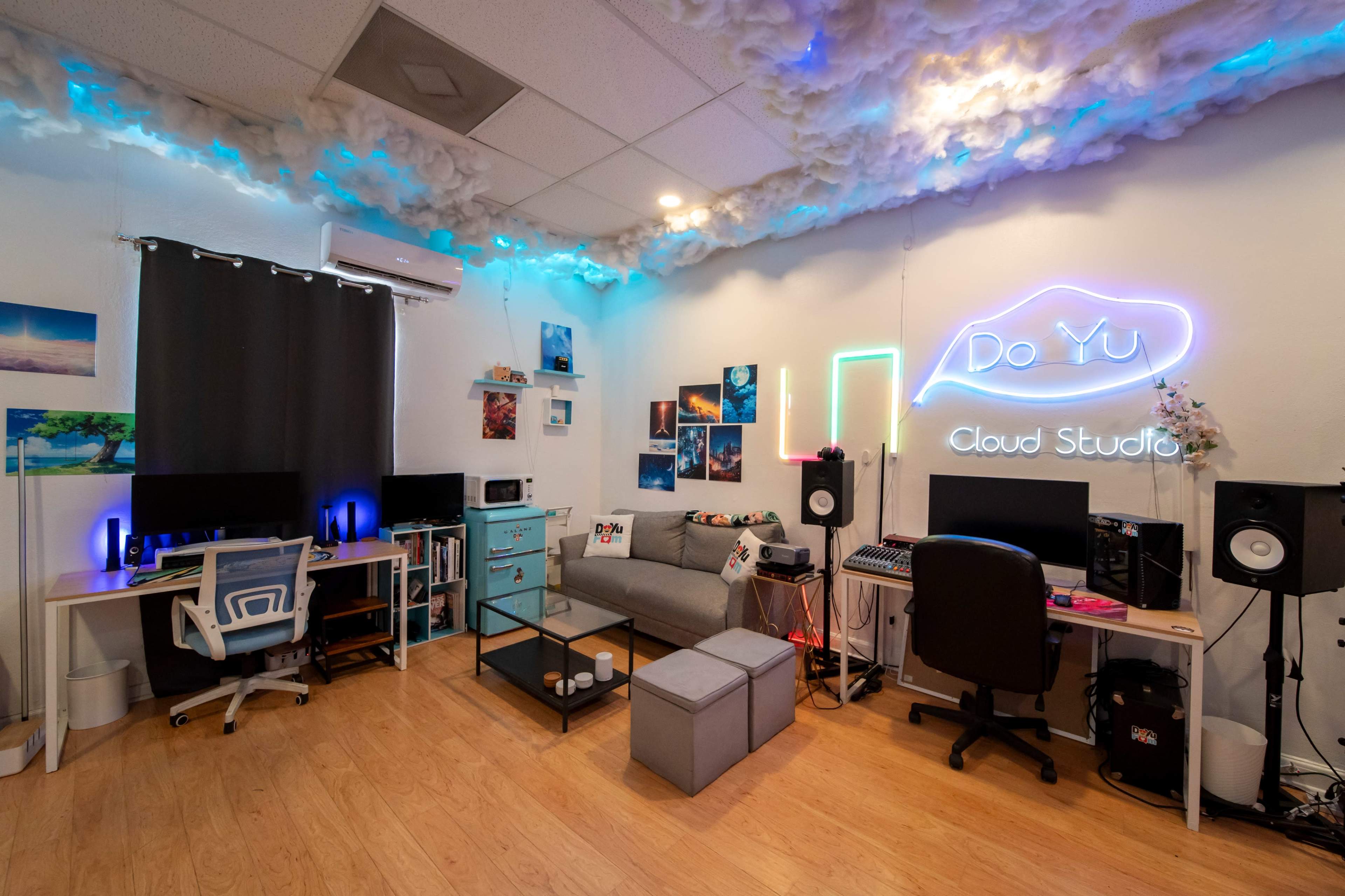 The image shows a modern studio space with two workstations, colorful wall decor, and cloud-like ceiling installations, illuminated by neon lights.