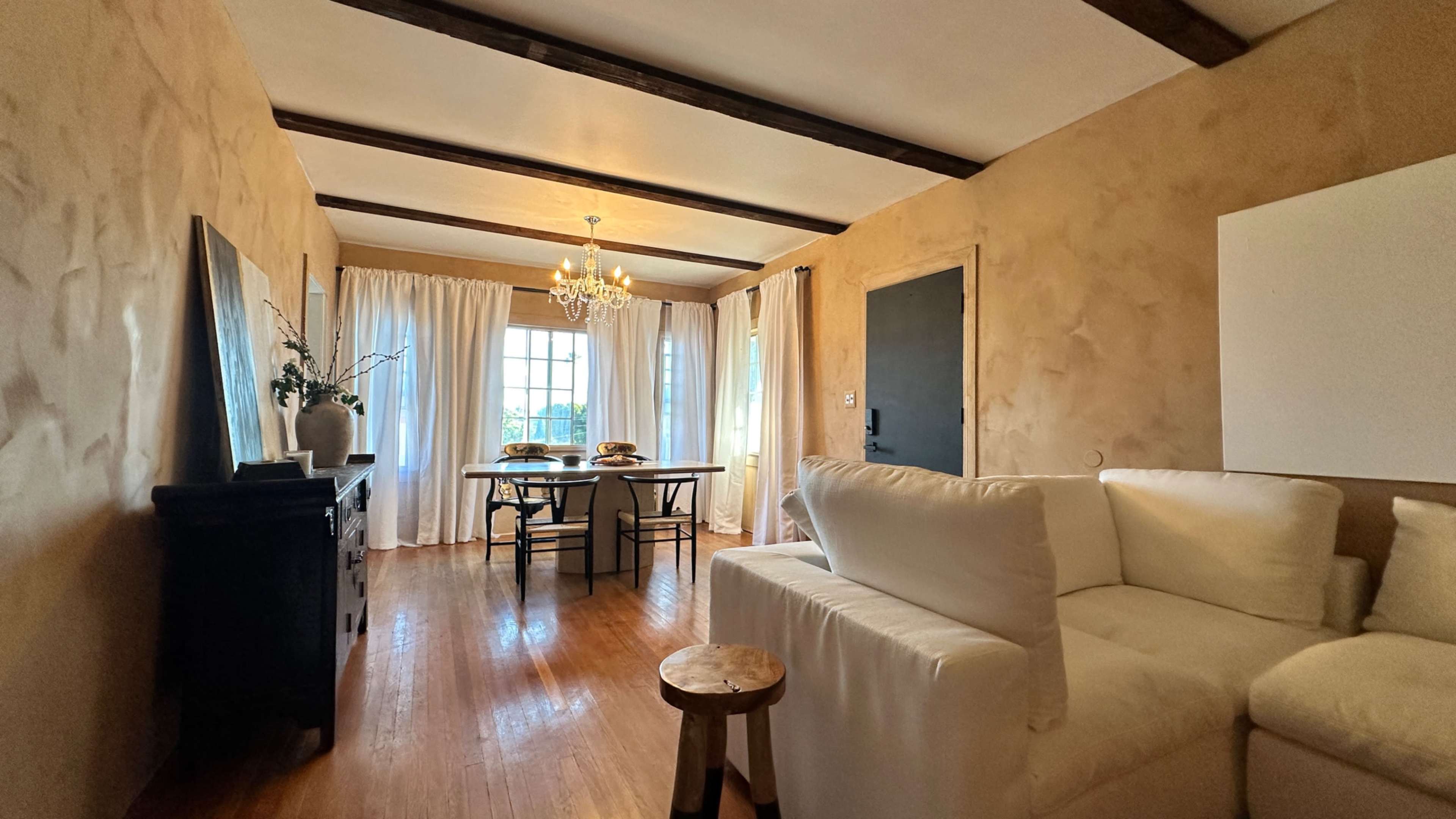 A living space with a white sofa, a wooden table set for dining, and large windows covered with light curtains.