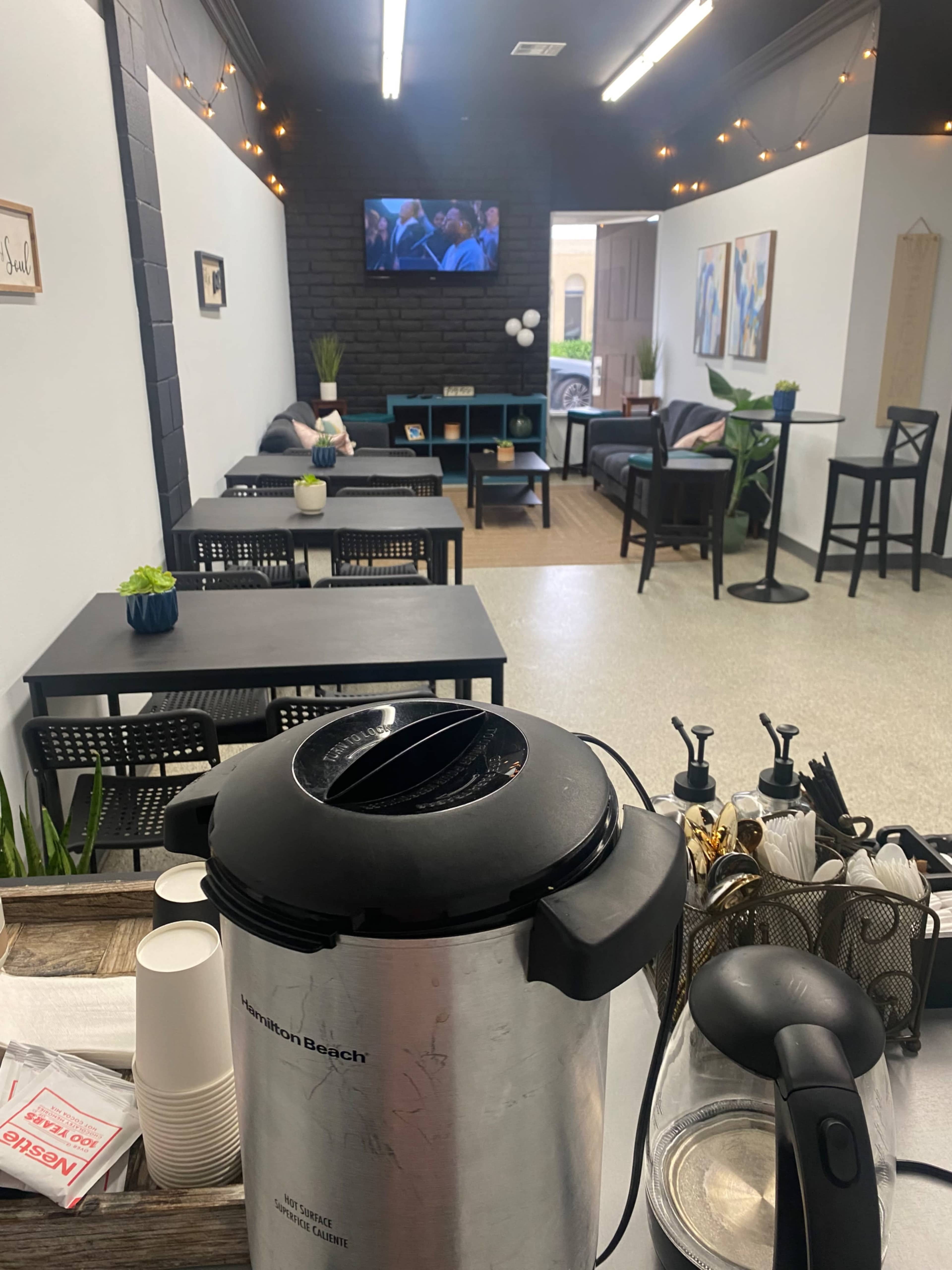 A modest indoor space with a coffee urn in the foreground, tables and chairs arranged for seating, and a television mounted on the wall.