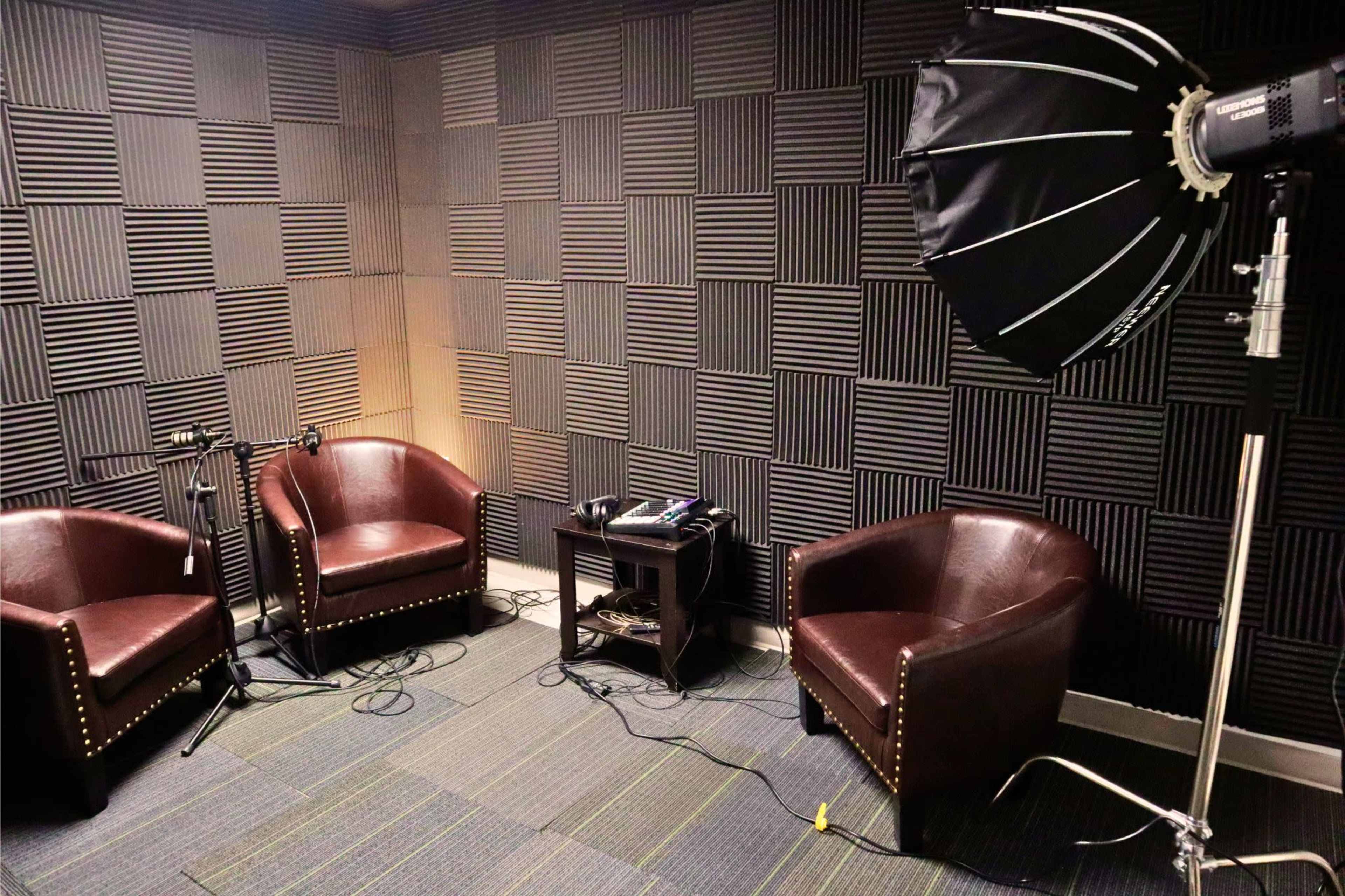 The image shows a soundproof room with two brown leather chairs, a small table, and professional lighting equipment set up in the corner.