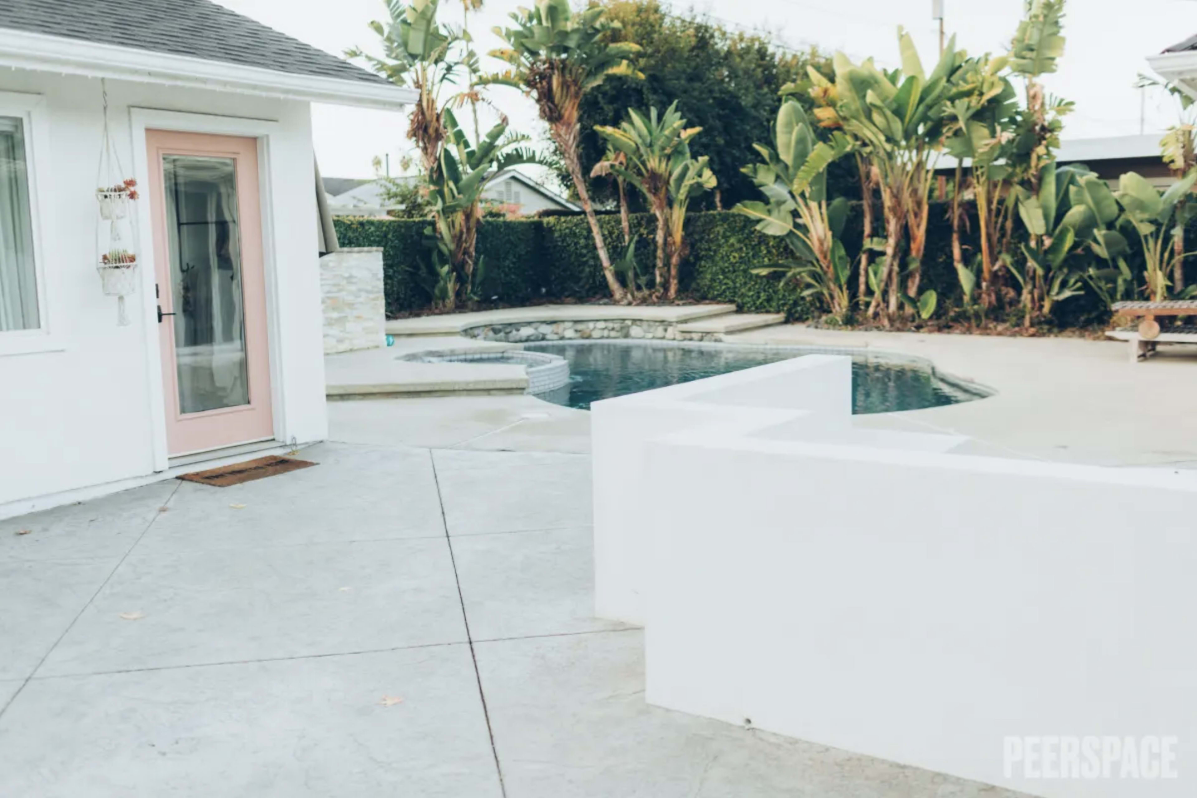 The image shows a swimming pool area enclosed by tropical plants and a white building with a pink door.
