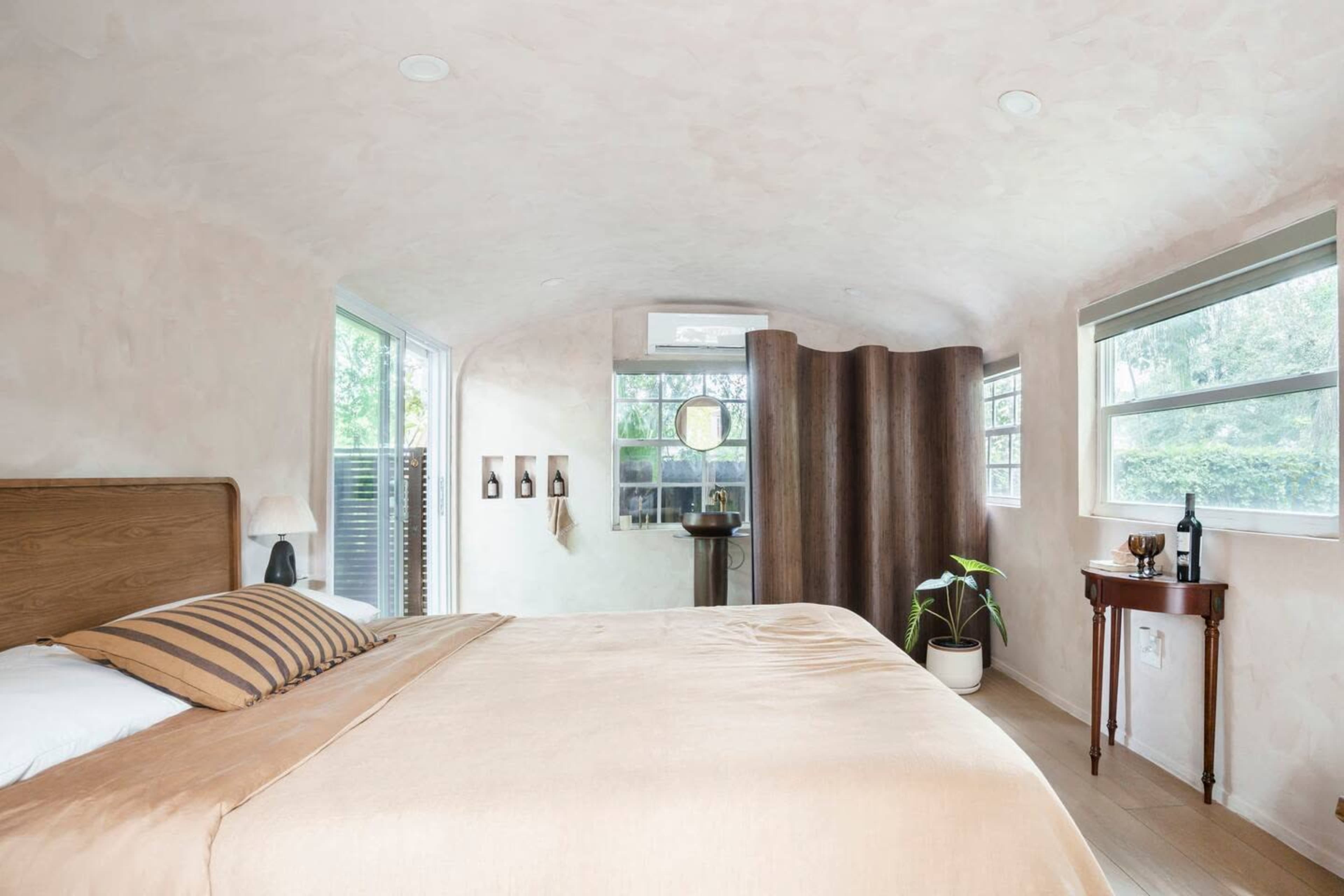 A softly lit bedroom with a curved wall, a bed with neutral linens, and a small side table next to a large window.