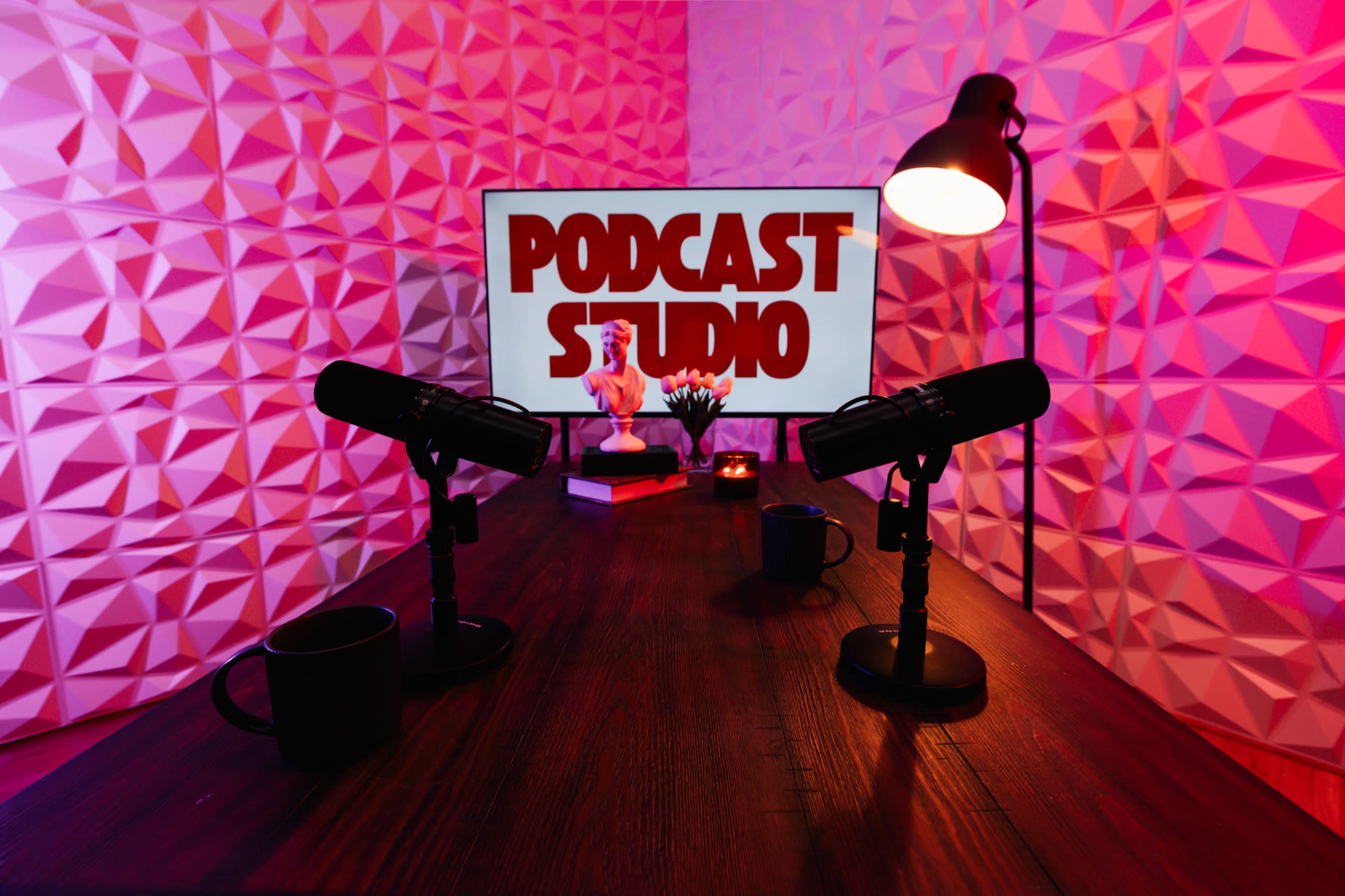 The image shows a podcast studio setup featuring two microphones on a wooden table, a screen displaying the text "PODCAST STUDIO," and ambient lighting.