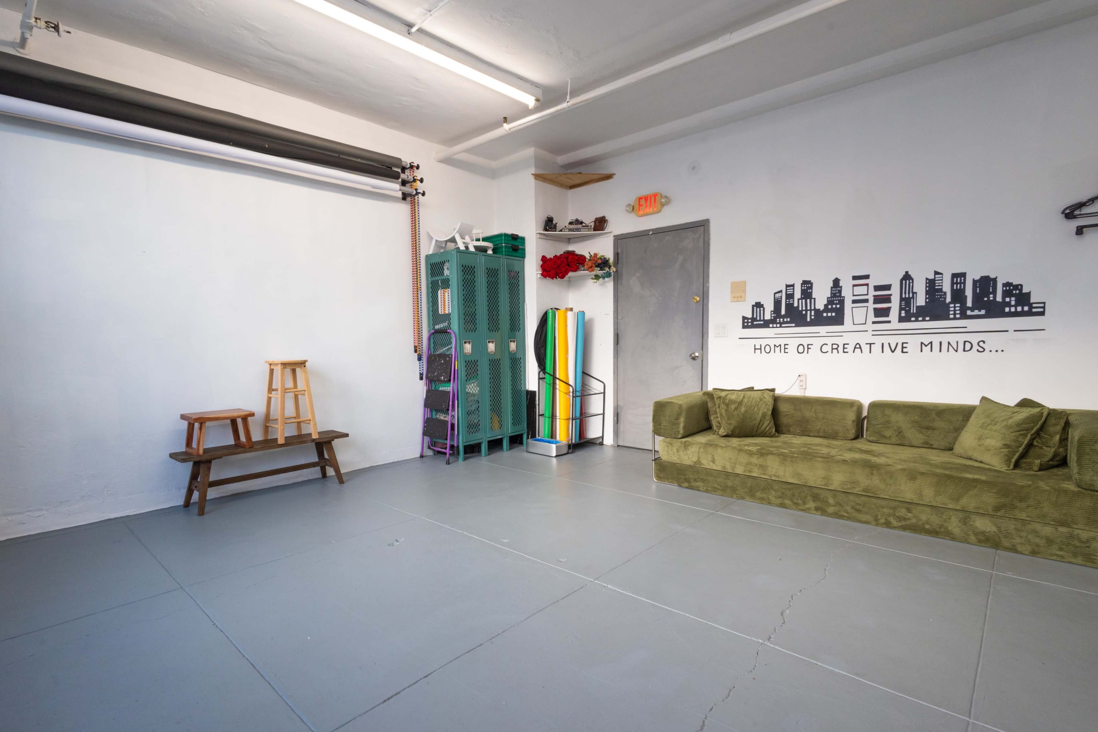 The image shows a spacious room featuring a green couch, a wooden stool, a set of lockers, and a wall decorated with a graphic of a city skyline.