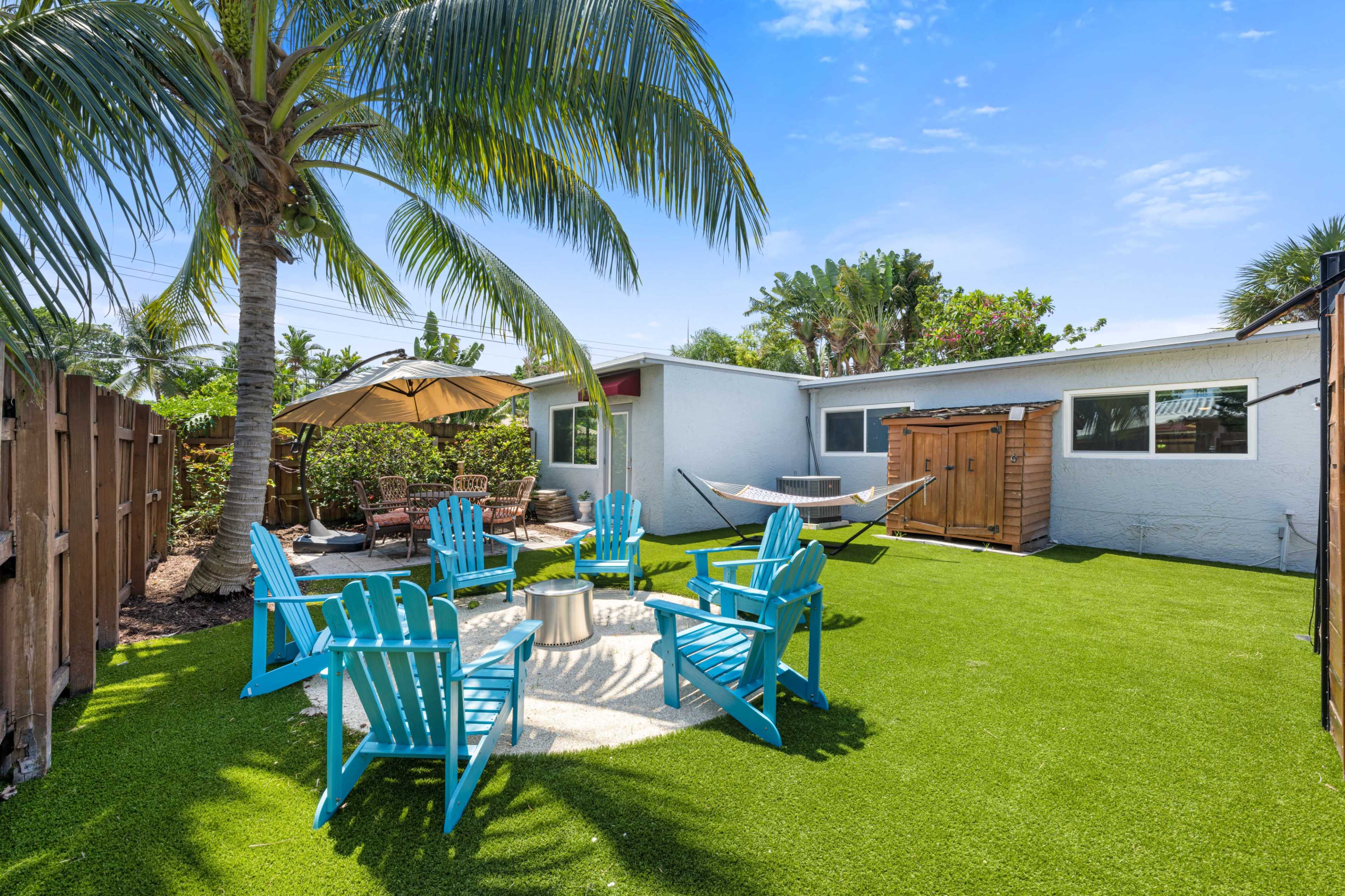 A backyard with artificial turf, featuring blue Adirondack chairs, a hammock, a shaded seating area, and a wooden shed.