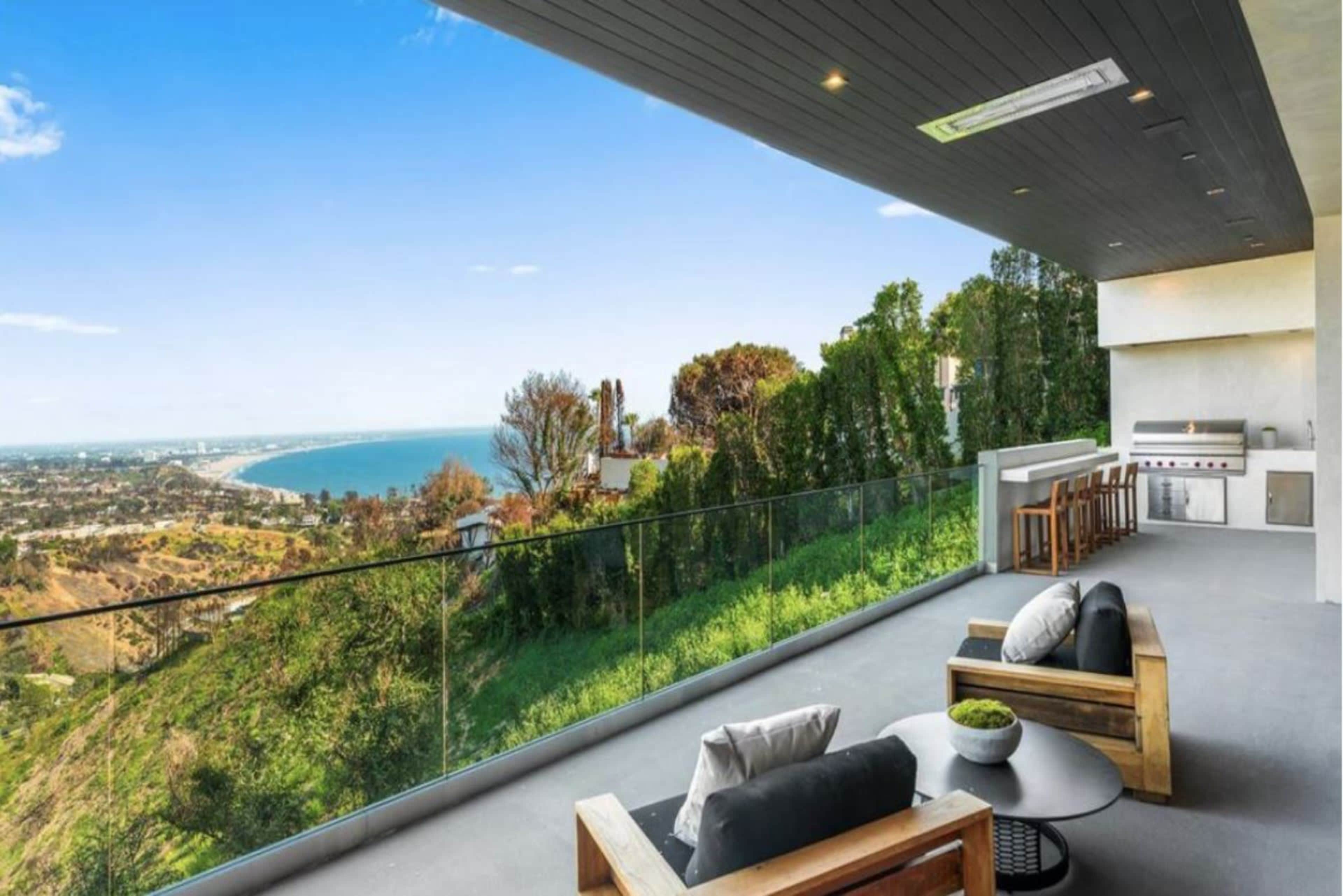 The image shows a spacious balcony with modern outdoor furniture overlooking a coastal view and greenery.