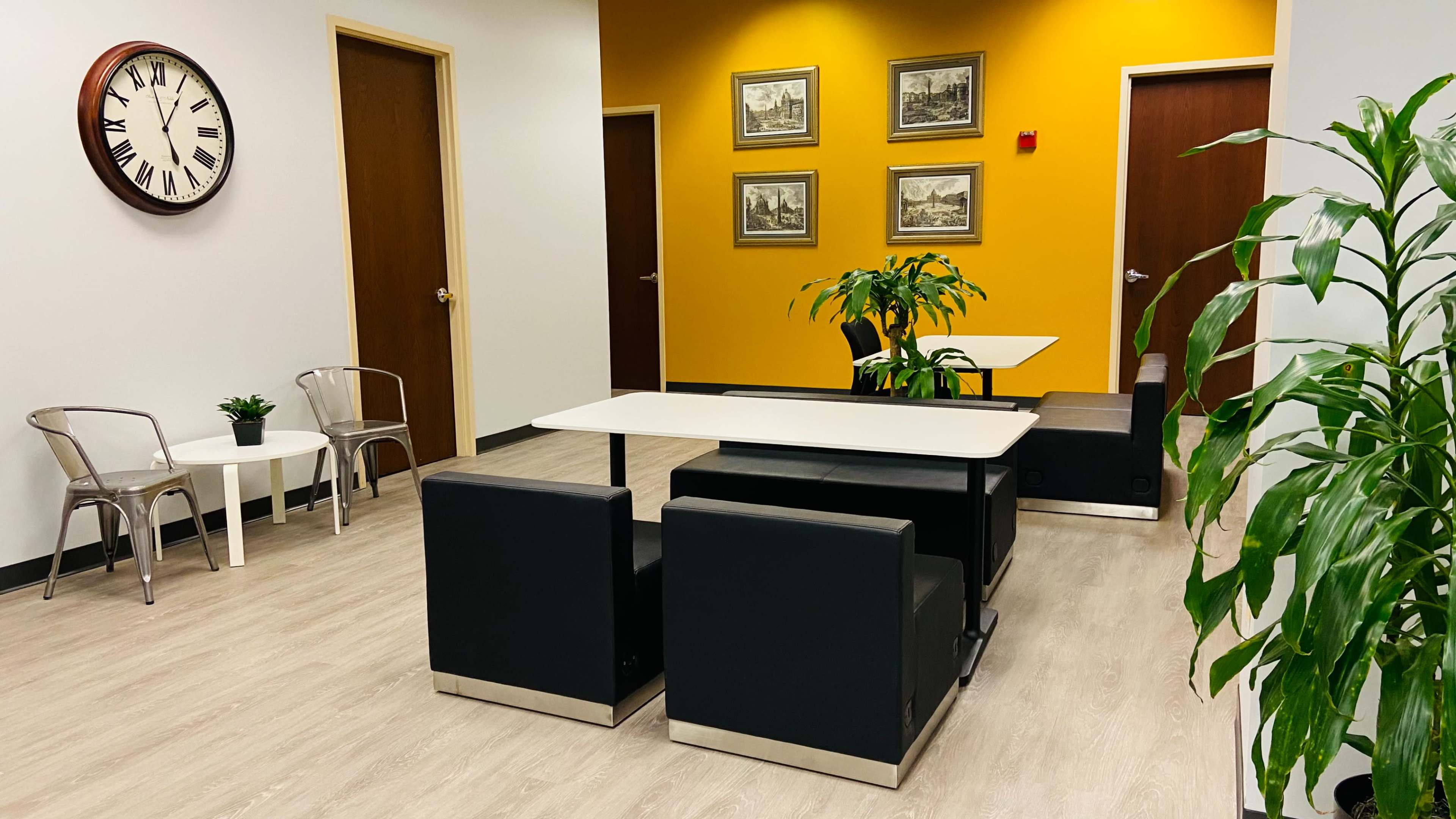 A modern lounge area with a clock on the wall, seating arrangements, a table, and potted plants against a backdrop of yellow and neutral-colored walls.