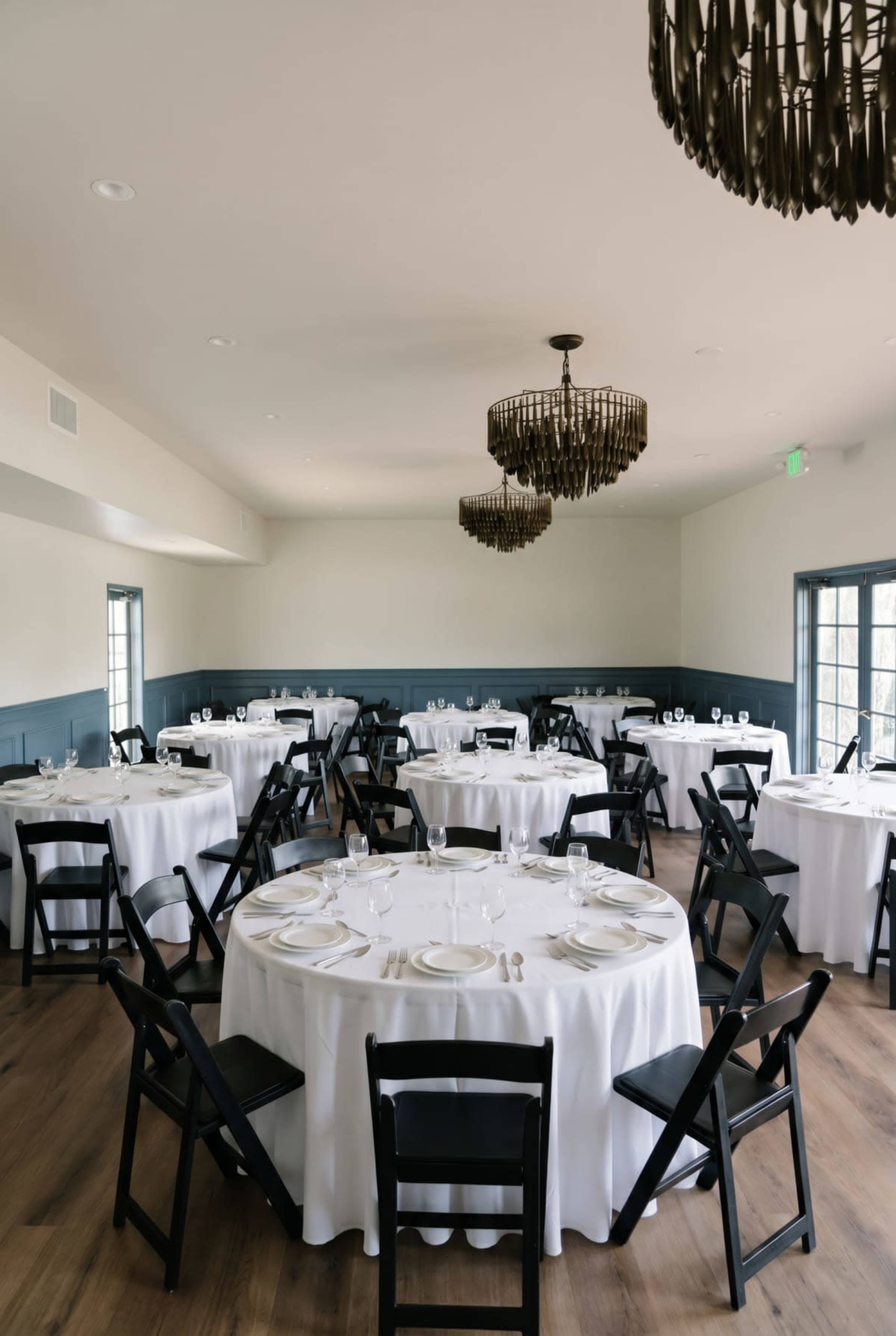 The image shows a dining room set up with several round tables covered in white tablecloths and black folding chairs arranged around them.