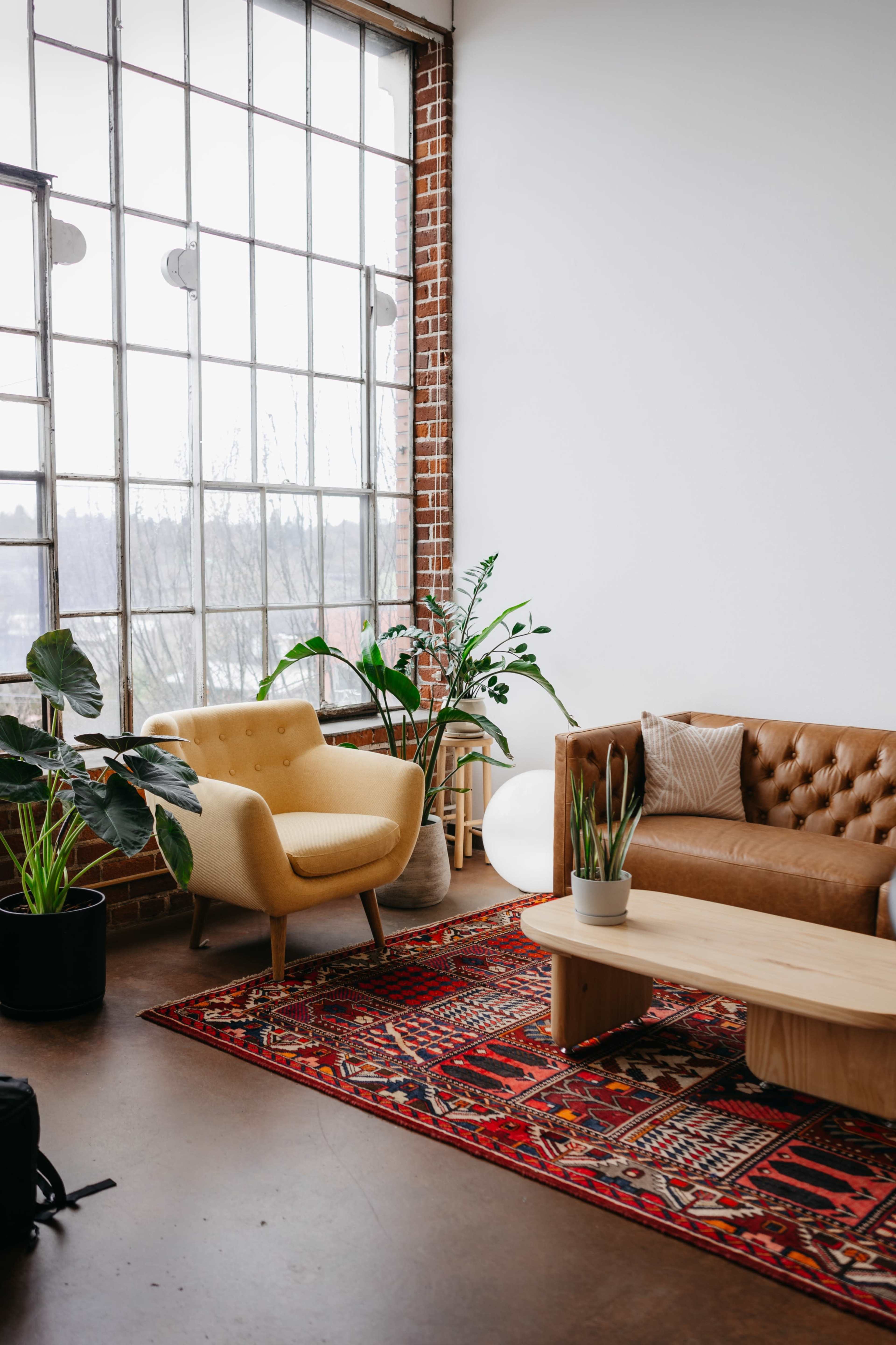 A cozy living room features a tan leather sofa, a yellow armchair, a wooden coffee table, a colorful rug, and large windows surrounded by plants.