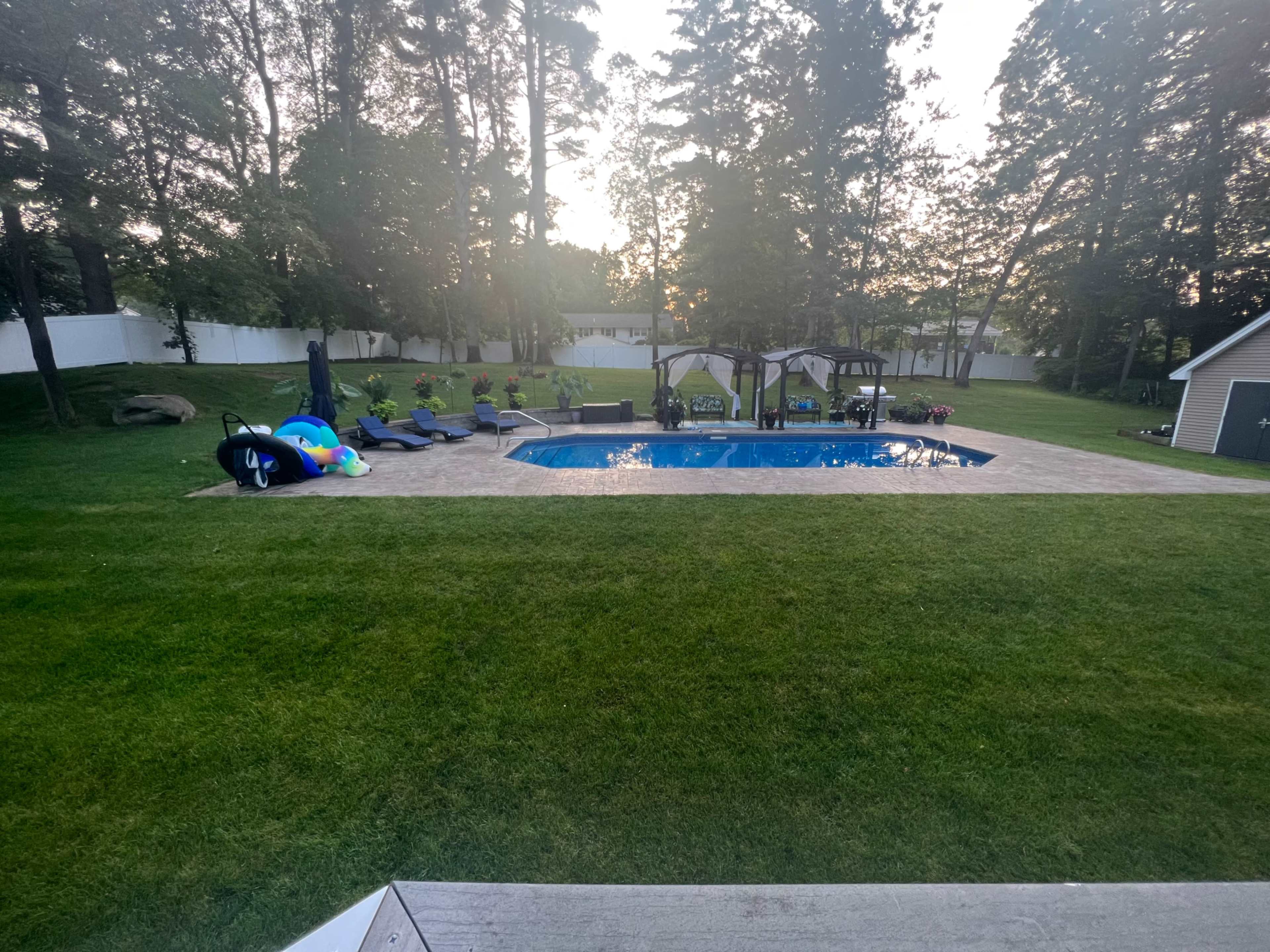 The image shows a swimming pool surrounded by lounge chairs and a gazebo in a grassy backyard with trees in the background.